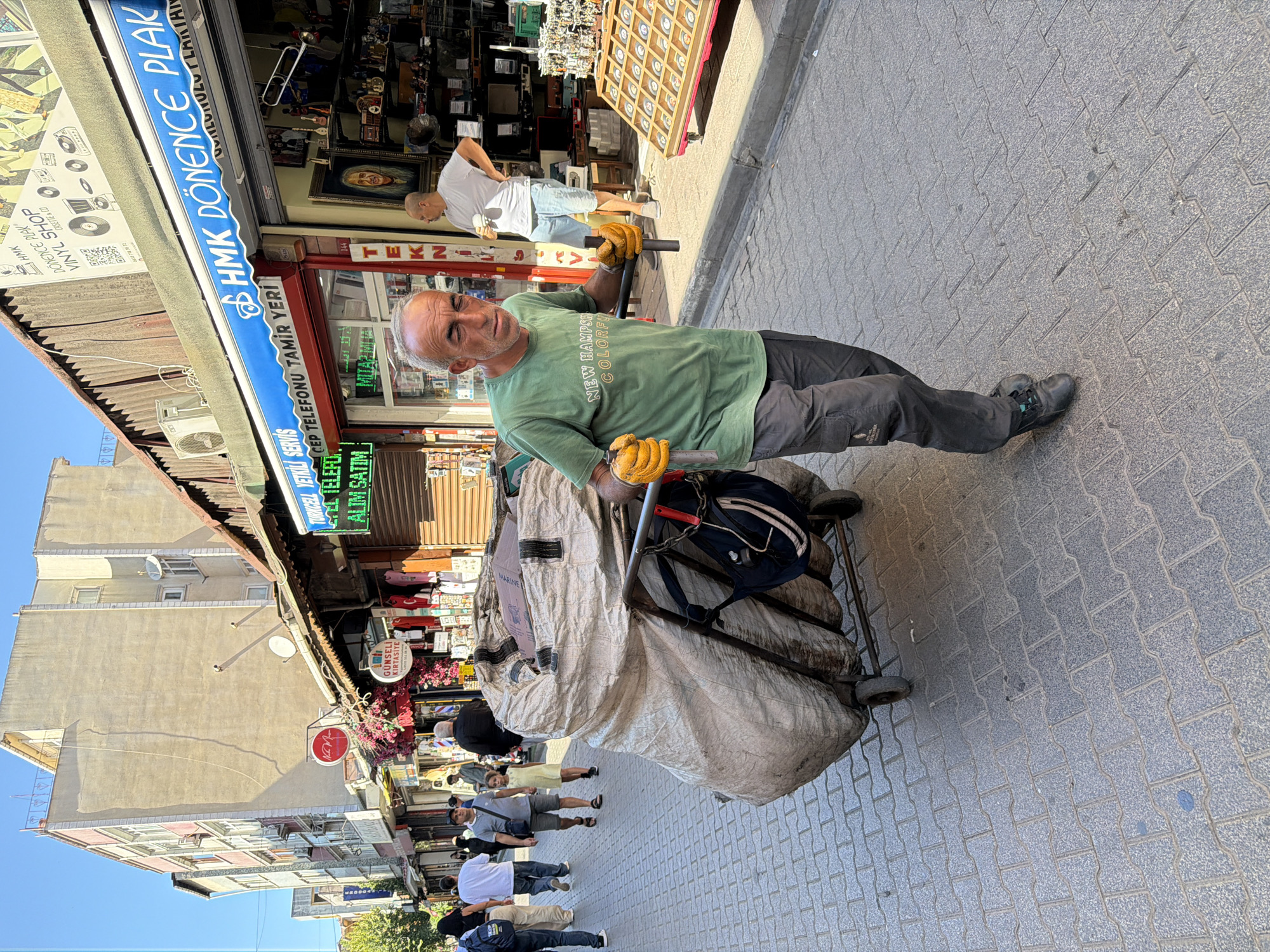 A man, wearing a green t-shirt and dark pants, pushes a large cart filled with bags down a cobblestone street in Istanbul, Turkey. He wears orange gloves and appears to be collecting or transporting the contents of the bags. Several shops line the street, with awnings extended over the sidewalks. People are walking in the background, some entering or leaving the shops. The architecture suggests an older urban environment. The lighting indicates it is daytime, with shadows cast on the street.