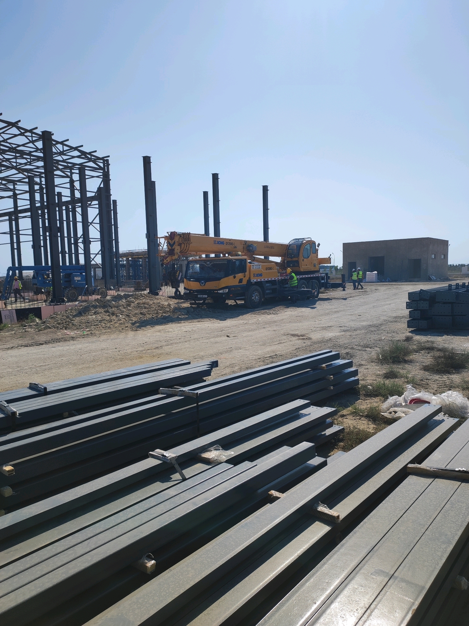 An outdoor construction scene in Khazar Raion, Azerbaijan, features a partially constructed steel frame building on the left. A yellow mobile crane with "XCMG" branding is positioned mid-frame, with its boom extended. Several workers, some wearing high-visibility vests and hard hats, are present near the crane and a small tan building in the background. A blue scissor lift is visible behind the steel structure. The foreground is dominated by stacks of grey metal beams and a dirt and gravel ground surface. The sky is clear and blue.