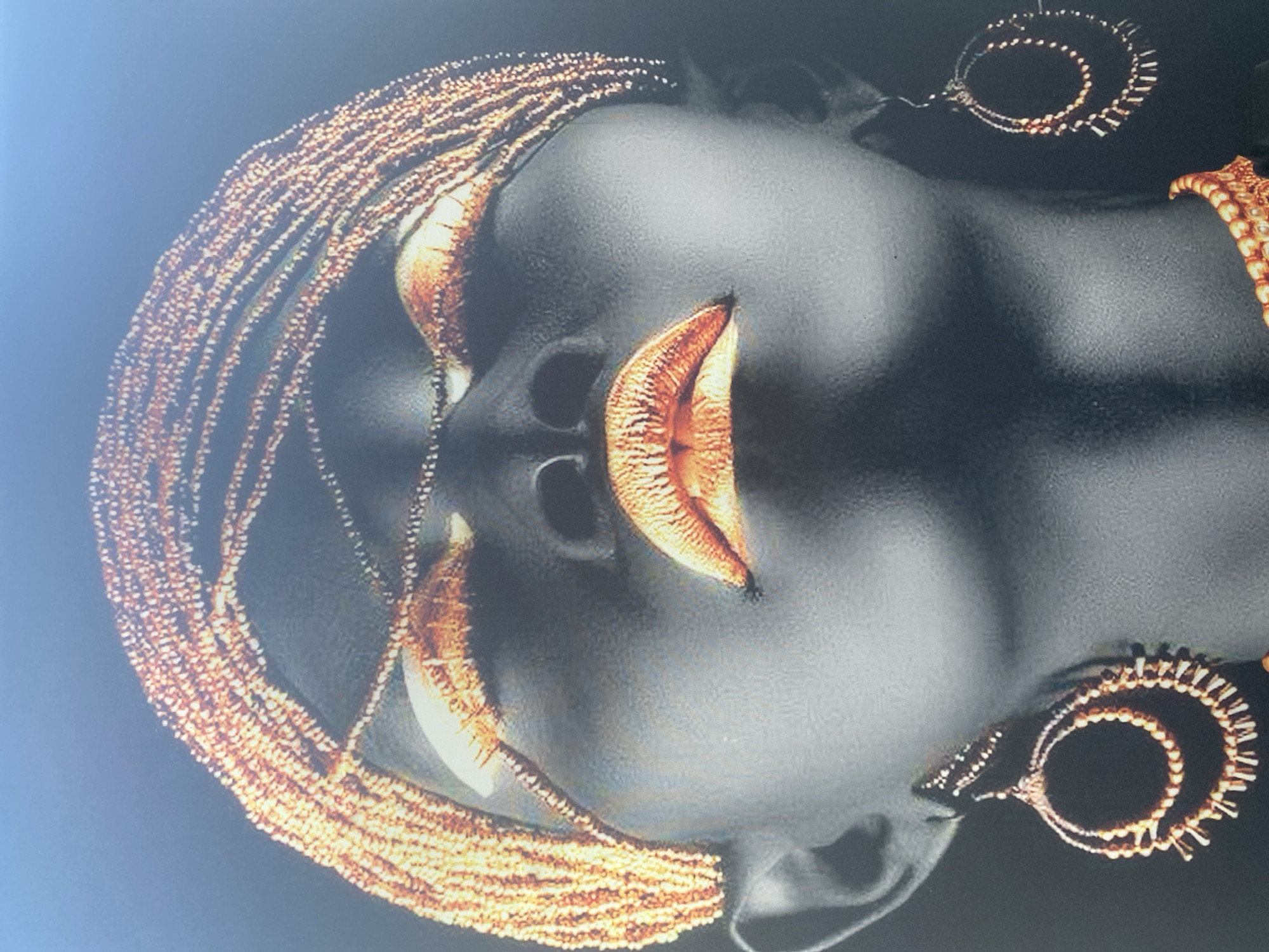 The image is a close-up, low-angle shot of a dark-skinned mannequin or statue adorned with beaded jewelry and makeup. The mannequin's face is tilted upwards, and its eyes are closed, with shimmering copper eyeshadow. Its lips are painted with a metallic copper lipstick that highlights their fullness. A prominent copper-beaded headdress encircles the mannequin's head, extending down to frame its face. Large, circular copper earrings with dangling beaded elements hang from its ears. A beaded necklace, also in copper tones, is visible at the bottom of the frame. The background is uniformly black, creating a dramatic contrast that emphasizes the subject. There is no discernible connection to Blagoevgrad, Bulgaria, in this image.