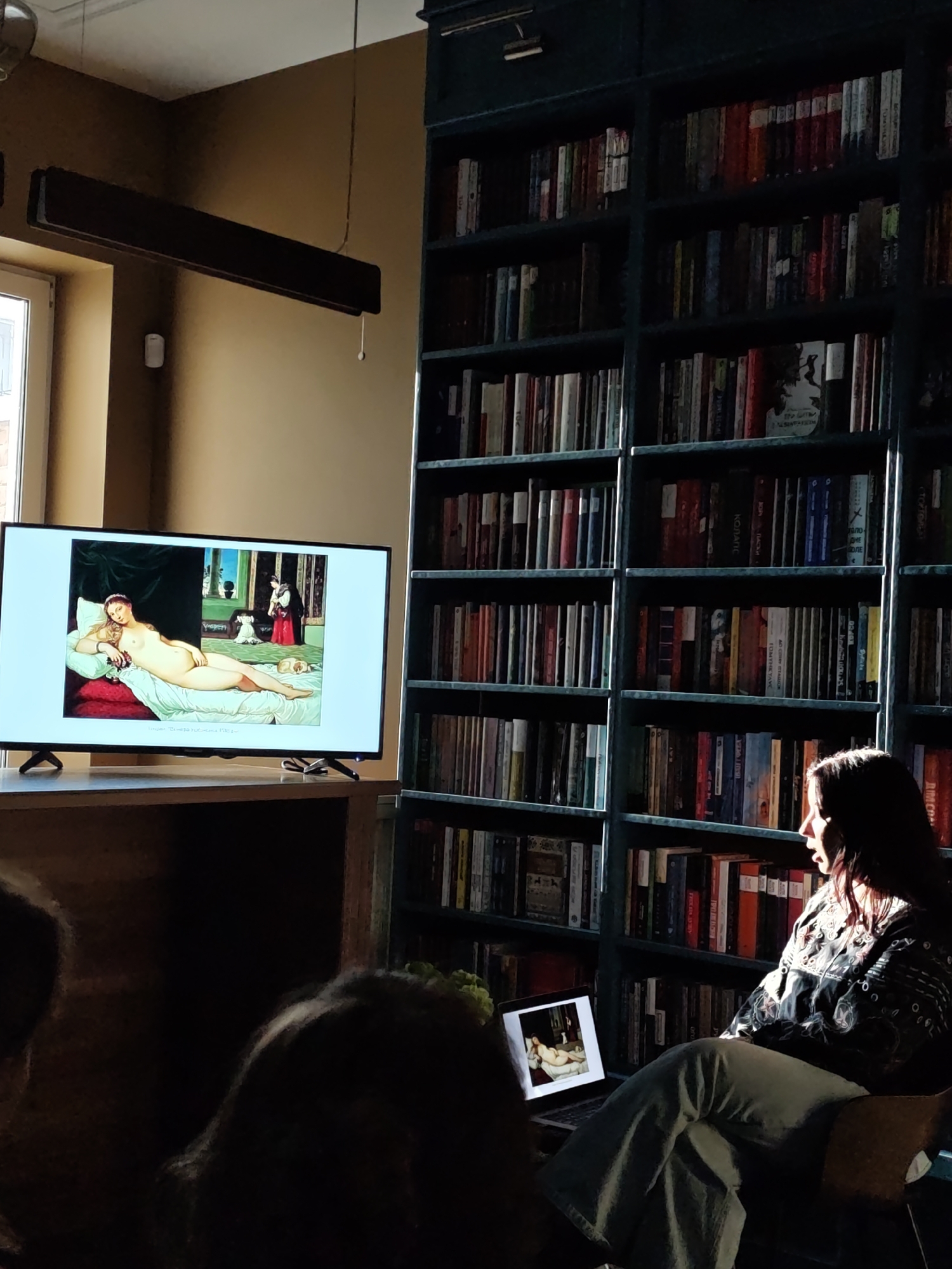 The image depicts an interior scene, likely a lecture or presentation taking place. A large screen displays a reproduction of Titian's "Venus of Urbino." Seated near the screen is a woman with dark hair, appearing to be watching the presentation, with a laptop displaying the same artwork. The background features a tall bookcase filled with books. While specific objects and visual references within the scene suggest cultural elements potentially associated with Vinnytsia, Ukraine, definitive confirmation of the city is not possible solely from the image.