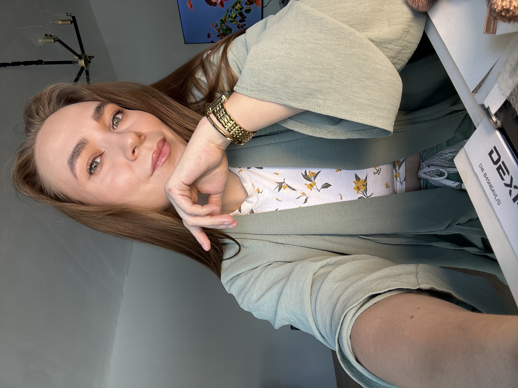 A woman with auburn hair and light complexion is posing indoors. She wears a light green blazer over a white floral top, and a gold watch. The background includes a television showing marine life, a modern chandelier, and a white desk with objects. The location in Kharnavy, Russia cannot be verified.