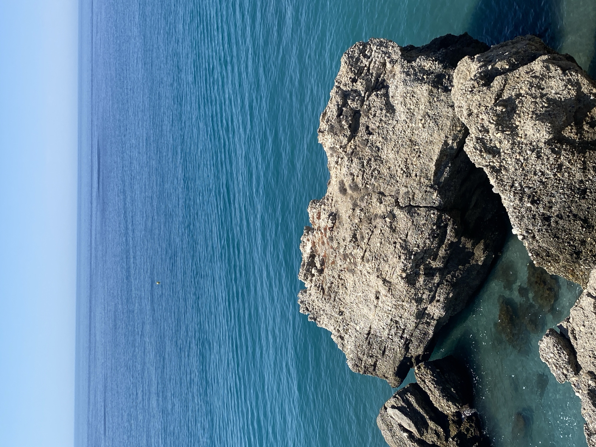 This is a high-angle, eye-level, medium shot of a rugged rocky outcrop jutting into the clear turquoise waters of the Mediterranean Sea. The sea stretches out to a horizon line under a bright, cloudless sky. A small yellow buoy is visible in the middle distance. The rocks are a light, weathered grey with visible textures and strata. The water is calm, with gentle ripples on its surface. The scene is characteristic of the coastline of Nerja, Spain, known for its dramatic rock formations and clear waters. There are no people or animals visible in the frame.
