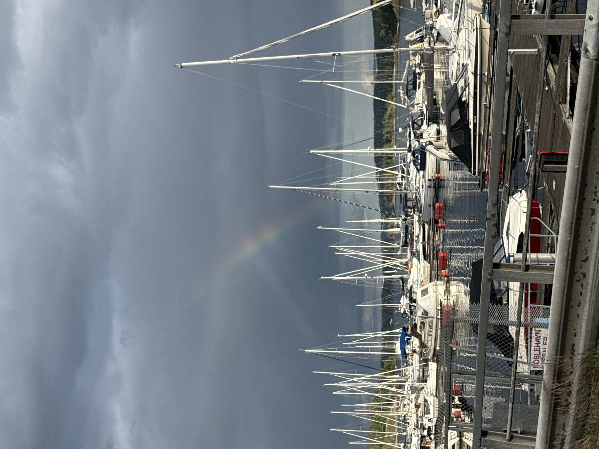 The image depicts a marina filled with sailboats and motorboats under a dramatic, cloudy sky with a rainbow arcing through it. The boats are docked, and the water appears calm. In the foreground, a fence and a railing suggest a viewing area or walkway. A sign on one of the boats or docks reads "KOBLEHAYN". The background shows a treeline and a body of water, characteristic of a coastal or fjord setting. The overall atmosphere is serene, with the rainbow adding a touch of natural wonder to the scene, which is identified as being in the city of Sandvika, Norway.