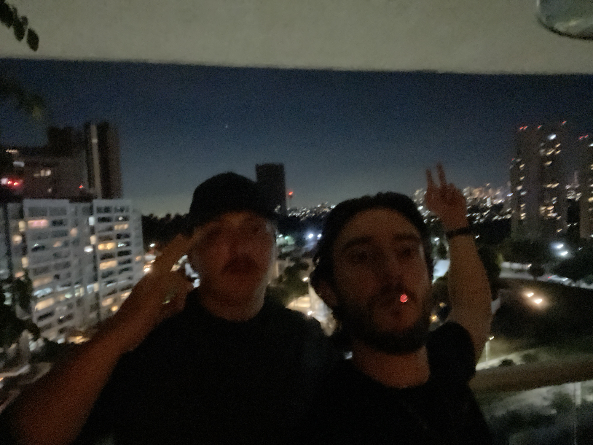 Two men are standing on a balcony at night in Huixquilucan de Degollado, Mexico, with a cityscape in the background. The man on the left is wearing a black cap and making a hand gesture near his head. The man on the right has a beard and is holding a cigarette in his mouth, while raising his other hand in a peace sign. Buildings with lit windows are visible in the background.