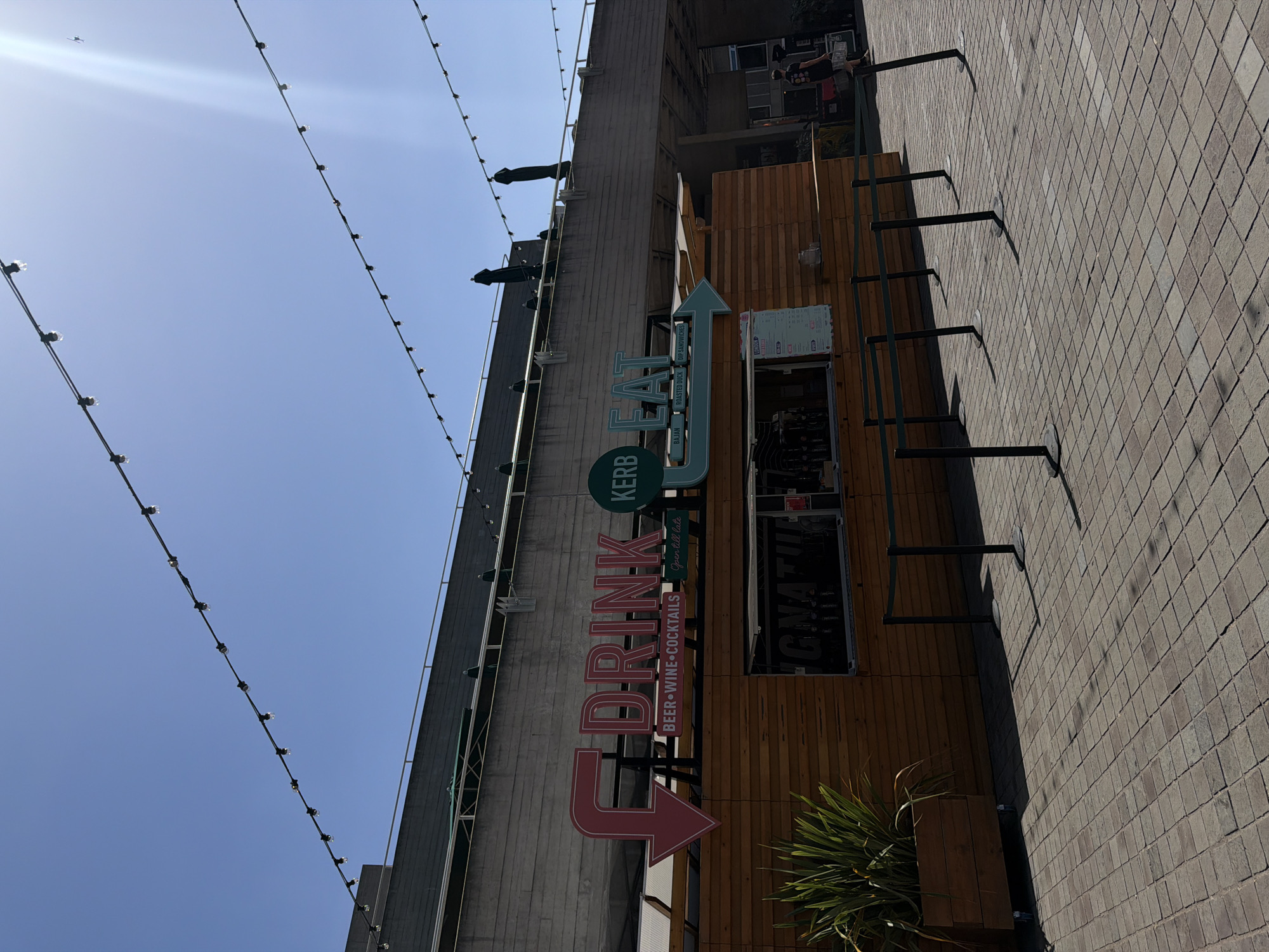 The image shows an outdoor shot of a KERB restaurant establishment, located in London, United Kingdom. The sign for the restaurant reads "DRINK" in red, with an arrow pointing downward, and "EAT" in blue, with an arrow pointing upward. The restaurant serves beer, wine, cocktails, and food such as roasted duck and DIP sandwiches. A string of lights is visible in the sky. The building housing the restaurant is tall and made of concrete. The wall adjacent to the restaurant is brick and has metal brackets.