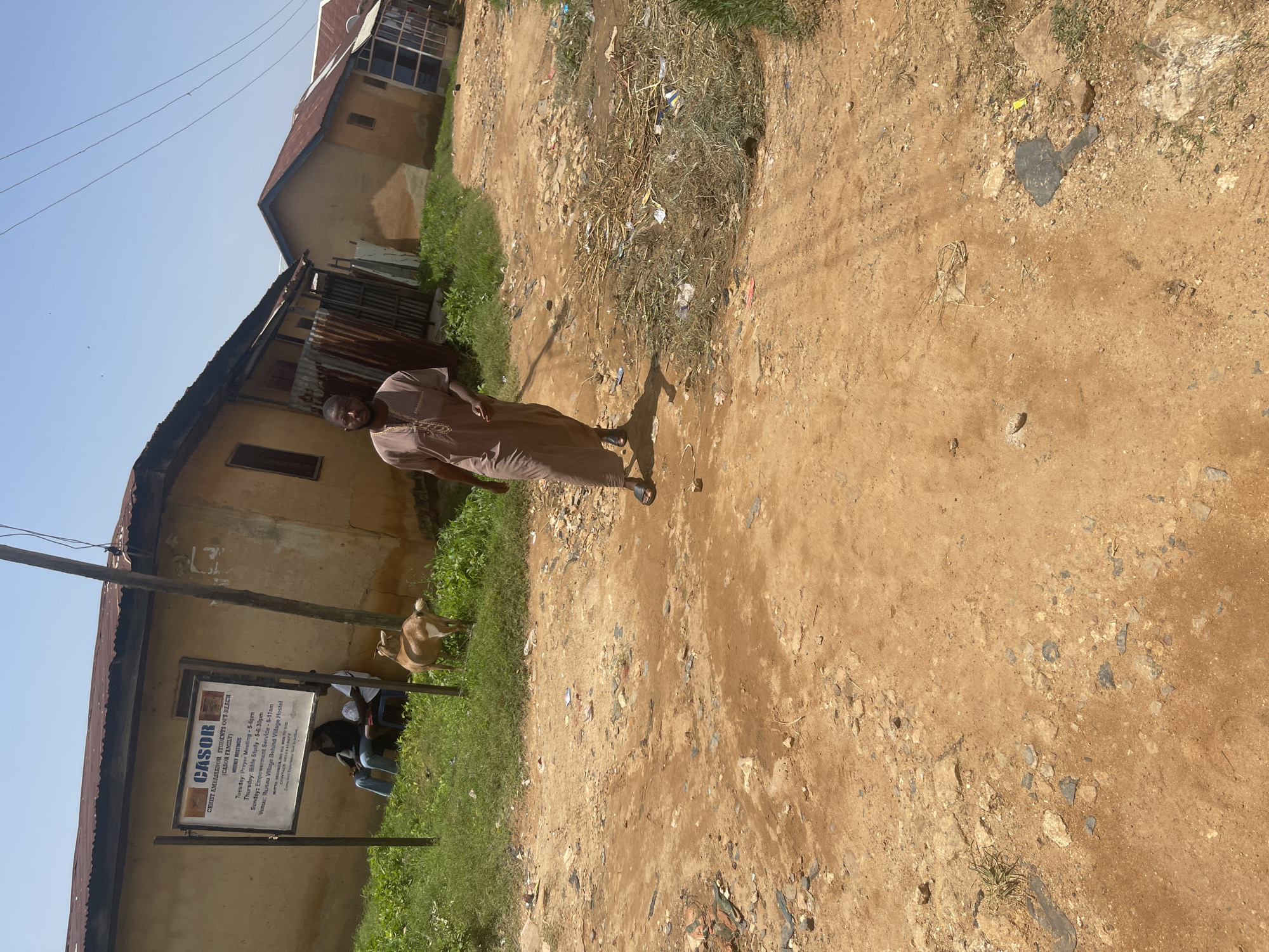 The image depicts a street scene in Jos, Nigeria, under a clear blue sky. A man in a brown, loose-fitting garment walks towards the camera on a dirt road strewn with debris and dry grass. To the left, a sign with text is mounted on poles in front of a building with a corrugated iron roof. Two individuals are seated on chairs beneath an overhang near the sign. A light brown goat stands on the grassy area to the left of the road, facing away from the camera. In the background, several buildings with similar architectural styles are visible, some with corrugated iron roofs and others with more modern window coverings. Electrical wires stretch across the upper portion of the frame.