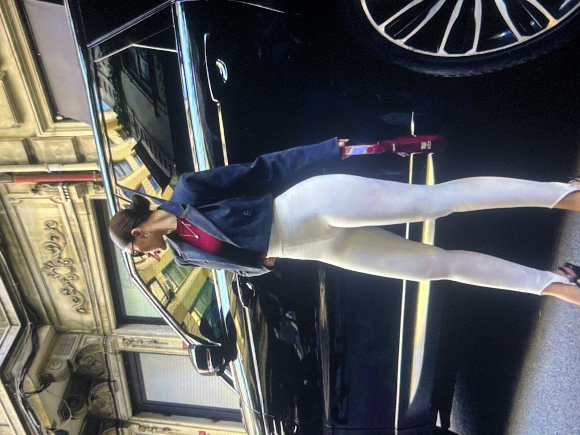 The image shows a person in white leggings and a dark jacket, standing next to a black car. The person is also wearing a red top and glasses. The car has a shiny black exterior and a large silver rimmed wheel is visible at the top right of the image. The background appears to be an urban setting with buildings reflected in the car's windows. The lighting suggests it could be daytime. The image is oriented vertically. There is no visible text.