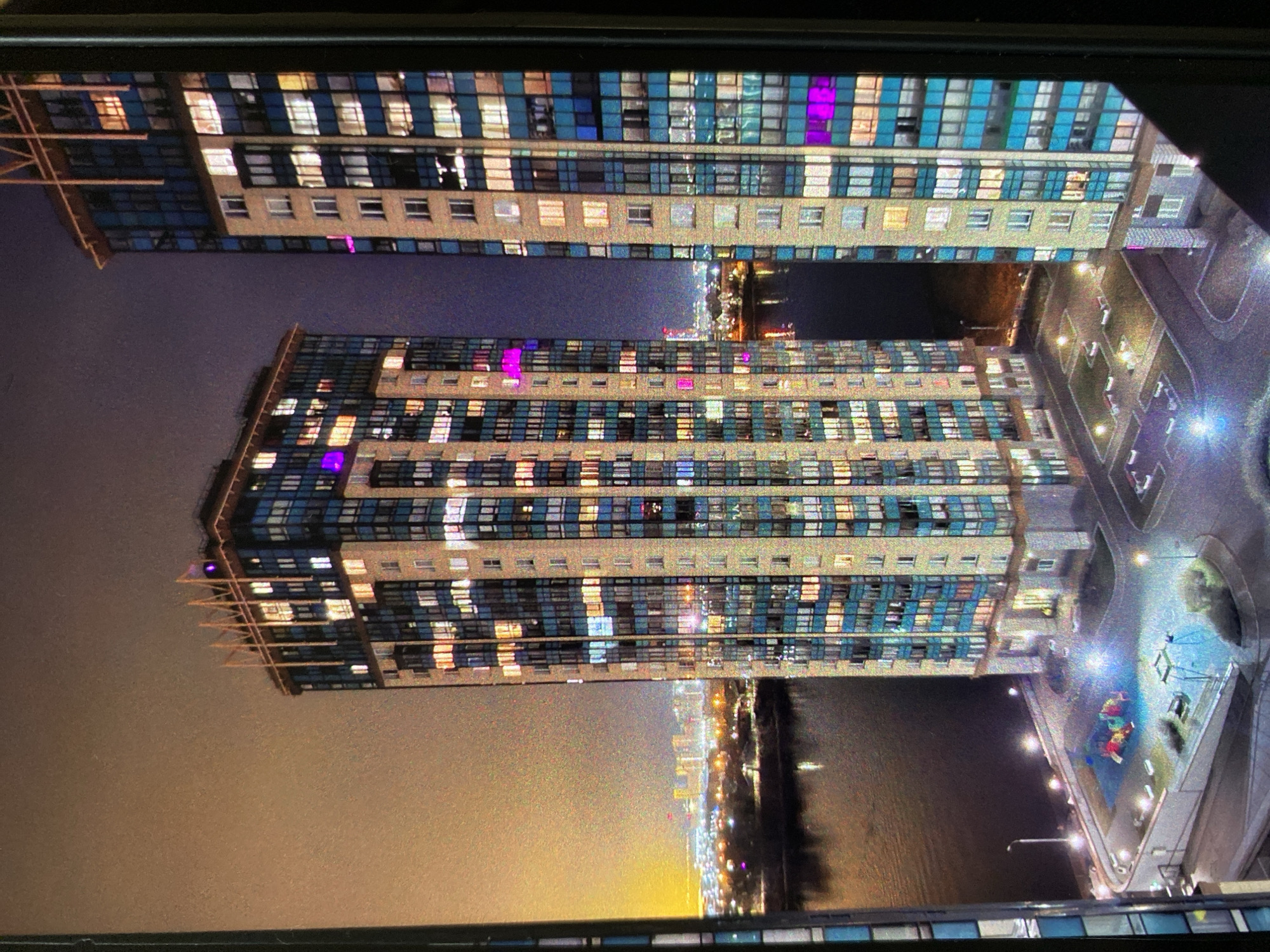 The image, oriented sideways, presents a nighttime urban landscape dominated by two modern high-rise buildings. These structures are detailed with numerous windows, many of which are illuminated in warm yellow, white, and distinct magenta or purple hues. Blue-tinted glass panels are also integrated into the building facades. One building exhibits visible structural elements, possibly architectural supports or scaffolding, extending from its side.
On the left side of the frame, a dark night sky is visible, subtly glowing over a distant cityscape on the horizon. To the right of the buildings, a developed ground area is illuminated by streetlights, revealing paved pathways and a playground with colorful equipment (red, yellow, blue elements). Beyond this ground level, a dark body of water reflects distant city lights, contributing to the vibrant urban panorama.
The scene captures the lively atmosphere of a city after dark. The image displays a grainy texture, suggesting it was taken in low light or is a photograph of a digital screen. No specific activities or people are discernible, and no visible text is present. This urban night scene is set in Saint Petersburg, Russia.