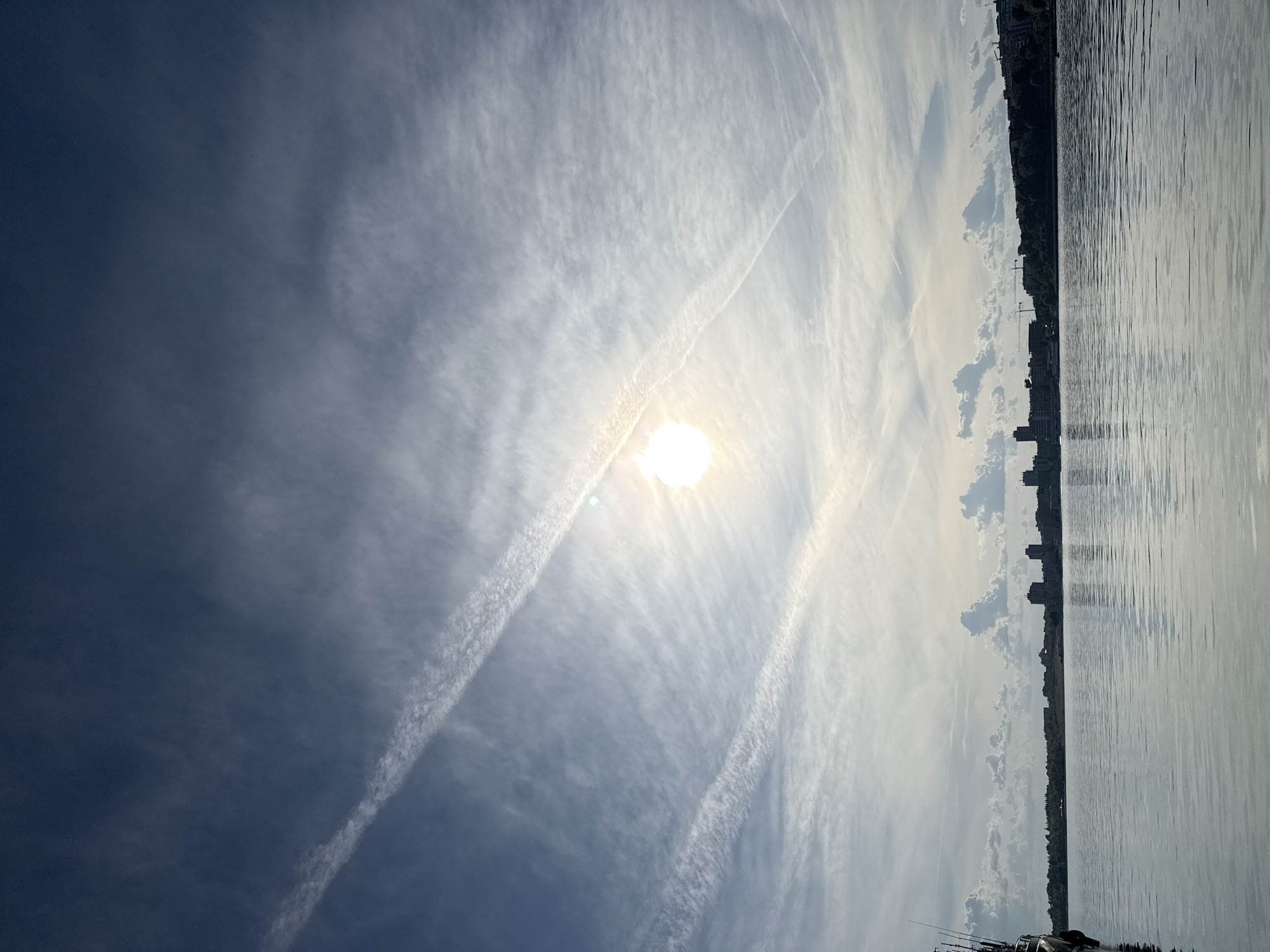 The image shows a scene likely taken at the beach in the city of Nesebar, Bulgaria. The view is slightly angled upwards towards the sky and oriented vertically. The sky is mostly blue with wispy clouds, including noticeable contrails from airplanes. The sun is visible, appearing as a bright white spot. On the right, a body of water stretches across the frame, with the shoreline visible in the distance. Buildings and trees line the coast. The lower portion of the picture includes a boat, or boats.