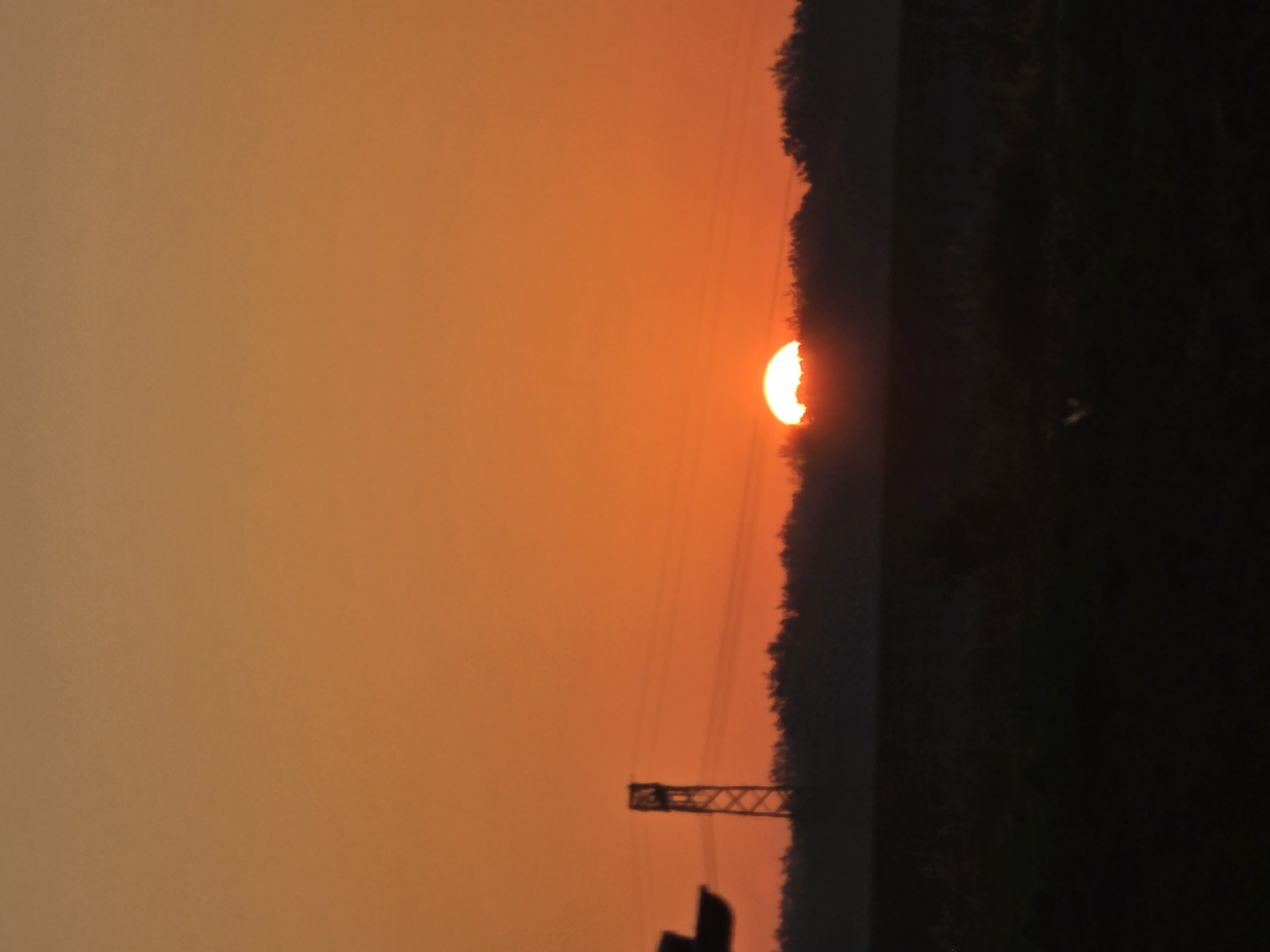 The image shows a sunset over a field in Kitzscher, Germany. The sun is partially obscured by a tree line, casting a warm orange glow across the sky. A silhouette of a power line tower is visible on the left side of the frame, with power lines stretching across the scene. The foreground is a dark, silhouetted field, likely agricultural land. The overall mood is peaceful and serene, capturing the beauty of a rural sunset.