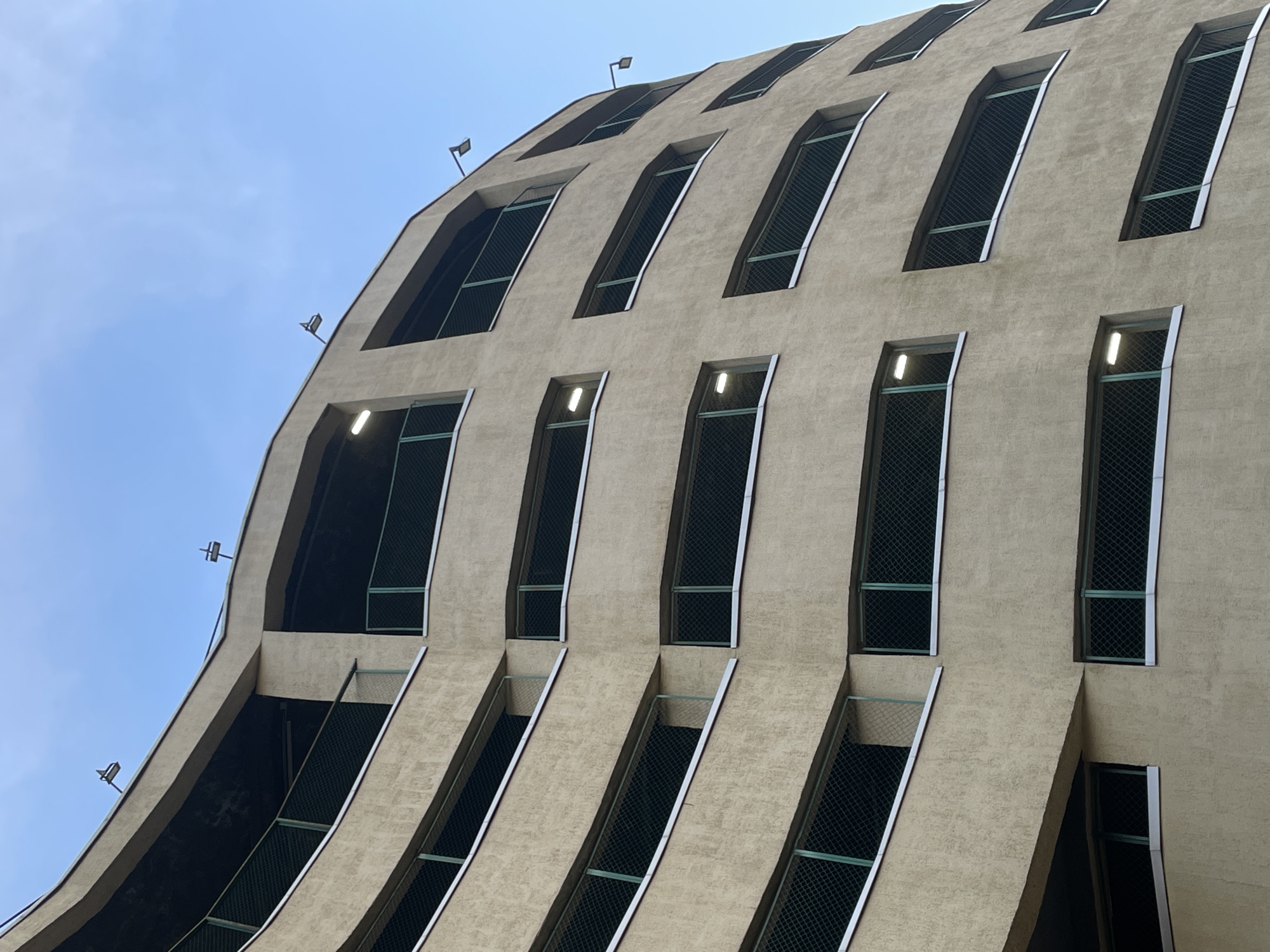 The image presents a low-angle, upward view of a modern building facade against a clear, light blue sky. The building is constructed from a light beige or tan-colored concrete or stucco, characterized by a distinctively undulating or wavy structure. The facade is punctuated by multiple rows of tall, narrow, vertically oriented openings.
These openings vary in their precise shape; some feature subtly curved or arched tops and bottoms, while others are more rectilinear. Each opening is covered by a dark, fine mesh or grid, suggesting they might serve as ventilation shafts or protected window-like elements rather than conventional glass windows. Several of these openings, particularly on the lower visible levels, are illuminated from within or at their upper edge by bright, white, rectangular light fixtures.
Small, dark-colored spotlights or security cameras are mounted at various points on the building's exterior, notably along its upper edges and corners, protruding slightly from the facade. The building material has a noticeable, slightly coarse texture. The clear, unblemished blue sky fills the upper left area of the frame, indicating a bright daytime setting. No people, specific activities, or visible text are present in the image. The architectural style is unique, offering no immediate clues to its geographic location.