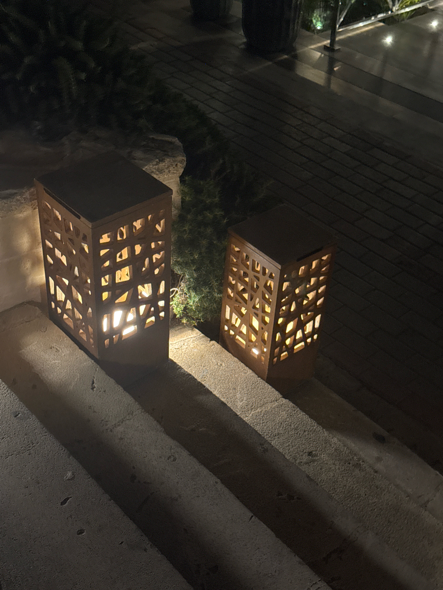 The image captures an outdoor scene at night, likely a pathway or staircase leading to an entrance in Agios Tychona Community, Cyprus. Two rectangular, dark-colored lanterns with intricate geometric cutouts are prominently featured. These lanterns are illuminated from within, casting warm, patterned light onto the surrounding concrete stairs and a patch of dense, dark greenery. The stairs are textured and appear to be made of stone, with distinct riser and tread details visible. The background is dimly lit, showing a paved walkway with faint reflections, possibly from overhead lighting or wet surfaces. The overall ambiance is one of subtle, atmospheric illumination, suggesting an inviting approach to a building or area at night.