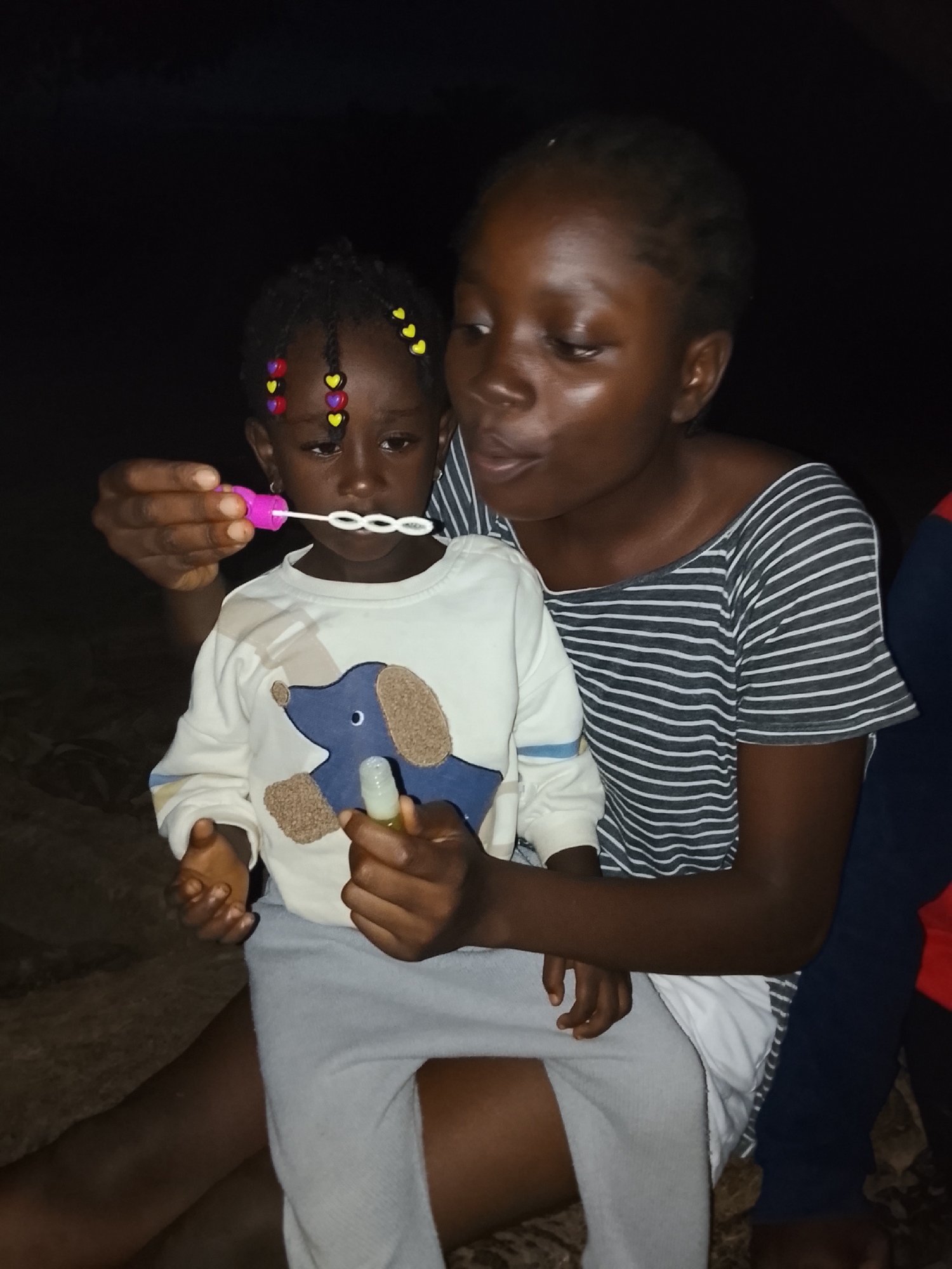 In this medium shot, two young Black girls are seated outdoors at dusk, likely in Akwanga, Nigeria. The older girl, wearing a grey and white striped off-the-shoulder top, is holding the younger girl on her lap. The younger girl, adorned with colorful heart-shaped beads in her braided hair, is wearing a white long-sleeved shirt featuring a blue dog graphic with brown paw prints. She is holding a bubble wand to her mouth, attempting to blow bubbles, with several already formed and floating in front of her. The older girl is gazing at the younger one, her mouth slightly open as if she is about to blow as well. In the foreground, the younger girl's hands are visible, one holding the bubble wand and the other clutching a small, clear bottle, possibly containing bubble solution. The background is dark, suggesting the image was taken as night falls. The overall mood is one of playful innocence and familial warmth.