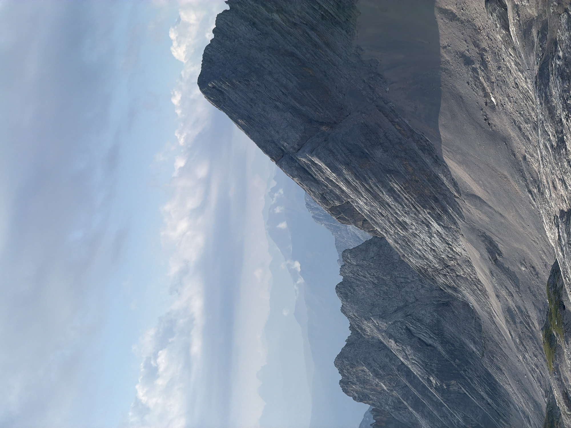 The image captures a majestic mountain landscape under a partly cloudy sky. In the foreground, a steep, rocky mountain slope dominates the right side of the frame, showing prominent layers and striations in the rock. To the left, a series of rugged, jagged peaks recede into the distance. The mid-ground features several layers of mountains, becoming progressively hazier and bluer, suggesting significant distance and atmospheric perspective. The sky is a light blue, with scattered white and gray clouds, some appearing wispy and others more solid. The overall scene depicts a high-altitude, alpine environment, likely within the Bavarian Alps. Without further information or context, specifying the exact location within Garmisch-Partenkirchen, Germany, is not possible from the image alone, though the terrain is consistent with the region's mountainous topography. There are no visible people or specific objects that indicate activity or human interaction in this particular frame.