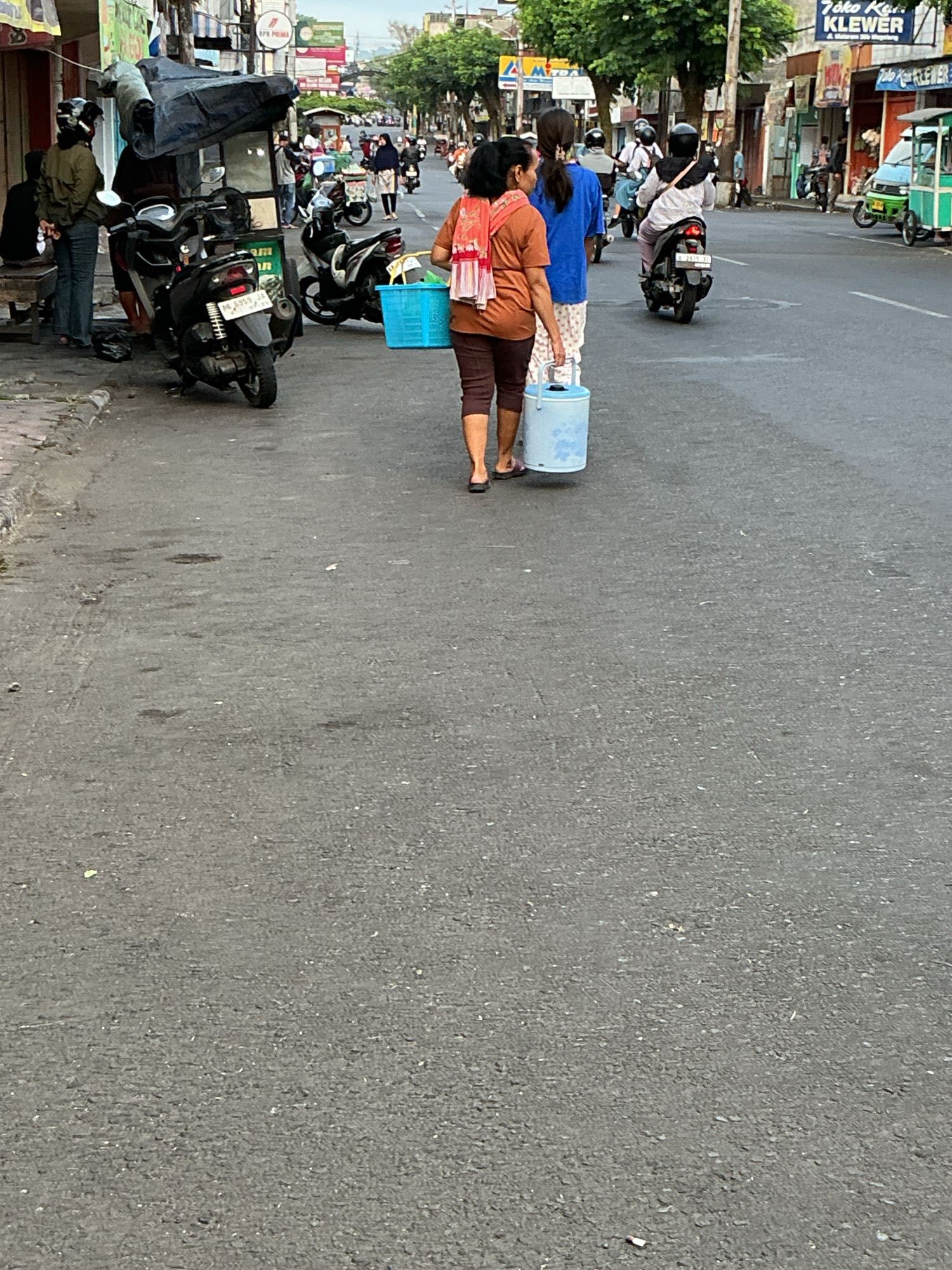 The image captures a street scene in Magelang, Indonesia. In the foreground, two women are walking away from the camera. The woman on the left is wearing a brown top and brown pants and is carrying a blue bucket. The woman on the right is wearing a blue top and patterned pants. Behind them, a person is riding a motorcycle. Several other motorcycles and a small green cart are visible on the street, along with buildings lining the road. The atmosphere appears to be that of a busy commercial area with ongoing daily activities.