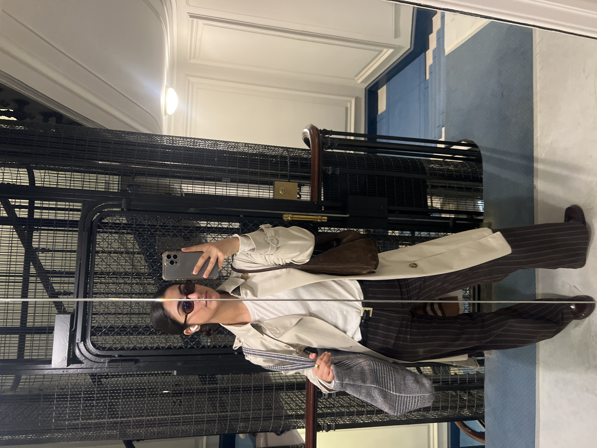 A reflection of a person standing in an elevator cage is captured in a mirror. The individual is wearing sunglasses, an earbud, a white shirt, striped pants, and a trench coat, and holding a handbag and phone. The interior of the elevator cage is visible, as is the wall behind the mirror, which features white panels and a blue-carpeted staircase. The scene appears to be within a building in Paris, France, as old-fashioned cage elevators are typical of buildings in this city.