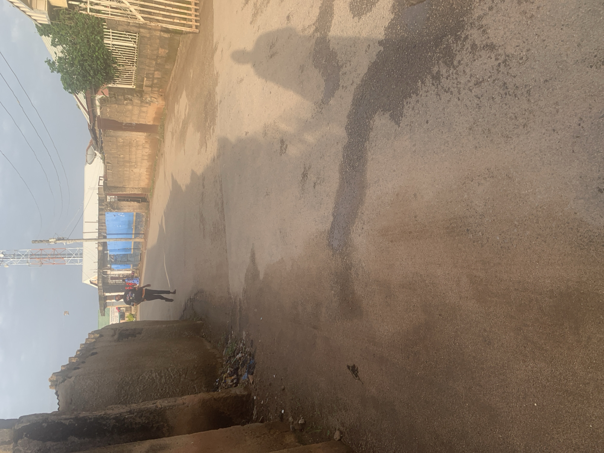 The image depicts a street scene in Jos, Nigeria. The road surface is asphalt, showing signs of dampness and discoloration, suggesting recent rain or a water spill. Shadows cast by the viewer and possibly a person are visible on the road, indicating it is daytime.
On the left side of the road, there is a low, rough-hewn wall, possibly part of a building or boundary. Behind this wall, a fence made of metal bars runs along the property line. Further down the street, a tall telecommunications tower is visible against a pale sky, suggesting an urban or semi-urban setting. There are also some structures with corrugated metal roofs and what appears to be blue tarpaulin sheeting.
In the middle distance, a young person, likely a male given their attire of dark clothing with a graphic print and dark trousers, is walking away from the camera. They appear to be the only clearly discernible person in the frame.
The overall impression is a slightly gritty, everyday street scene in Nigeria. The lighting and shadows suggest it is likely late morning or early afternoon. There is no visible text in the image.