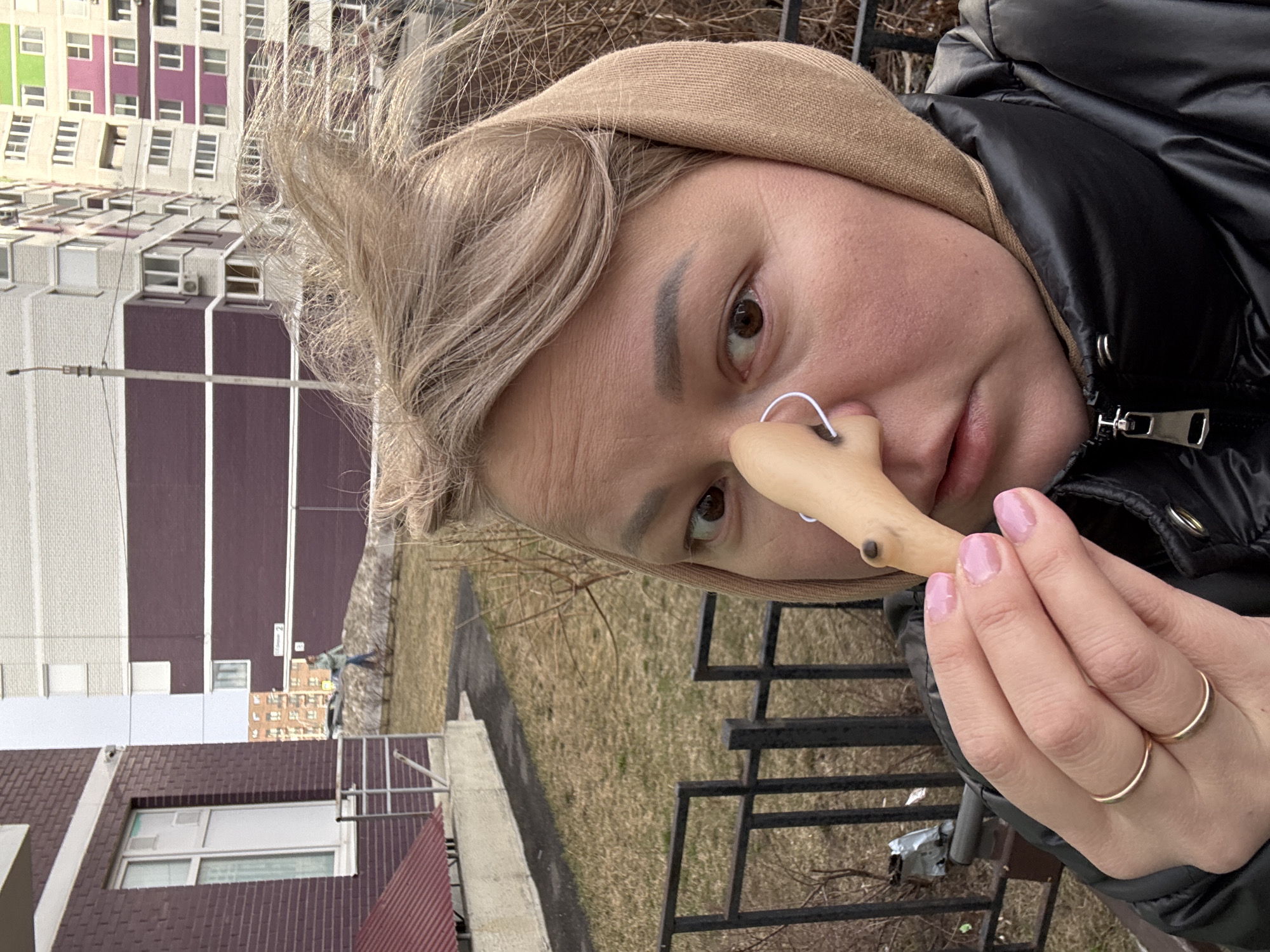 A woman in Izhevsk, Russia, is holding a wooden nose in front of her face. She is wearing a tan hood and a black jacket. The background includes a multi-story building with purple and white panels, and a patch of grass and bare trees. The woman has blond hair and two gold rings on her fingers. A thin white wire is wrapped around the wooden nose.