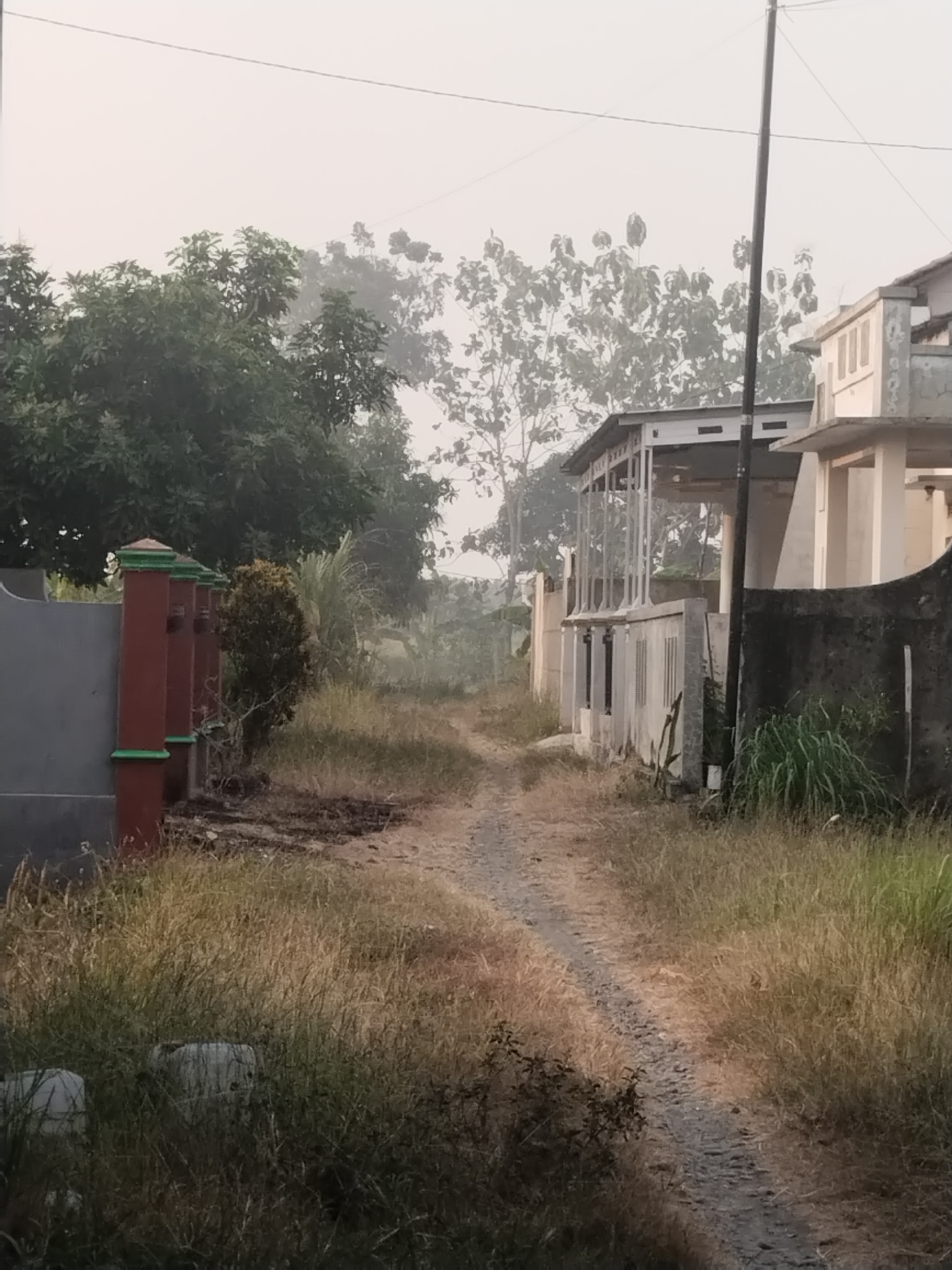 The image depicts a dirt path in Jembayat, Indonesia, leading towards a residential area. On the left side of the path, there is a grey wall with a decorative red and green post fence. Behind the fence, a dense green tree is visible. Further down the path, on the right, are several buildings. The closest building on the right is partially obscured by a dark, rough-textured wall. The building behind it has a white, two-story structure with columns and a balcony. The path is bordered by dry, tall grass and vegetation. In the background, trees and a hazy sky suggest a rural or semi-rural setting, possibly early morning or late afternoon due to the diffused light. There are no people visible in the image.