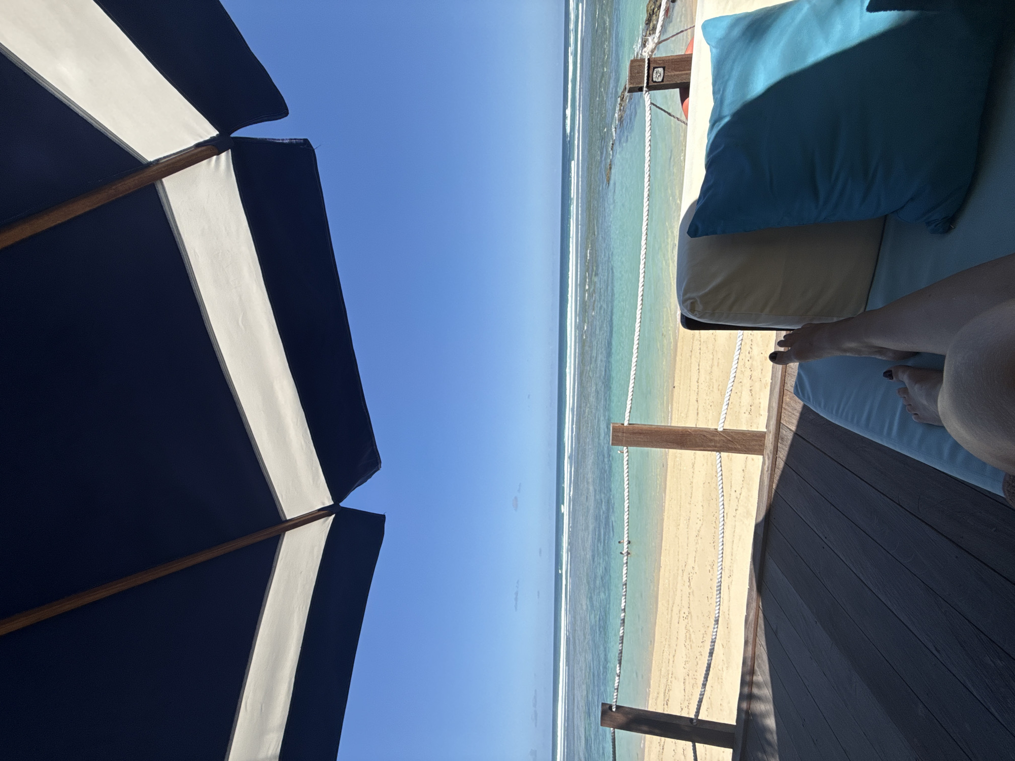 The image depicts an outdoor scene in Kuta Selatan, Indonesia, showing a beach view from a partially shaded lounge area. A blue and white striped beach umbrella dominates the left side of the frame, providing shade. To the right, the beach stretches out with clear turquoise water meeting the sandy shore. A rope fence is visible near the water’s edge. A lounge bed with blue pillows is on the right side. The lower right corner shows a person's legs and feet resting on the lounge bed. The sky is clear blue, indicating a sunny day.