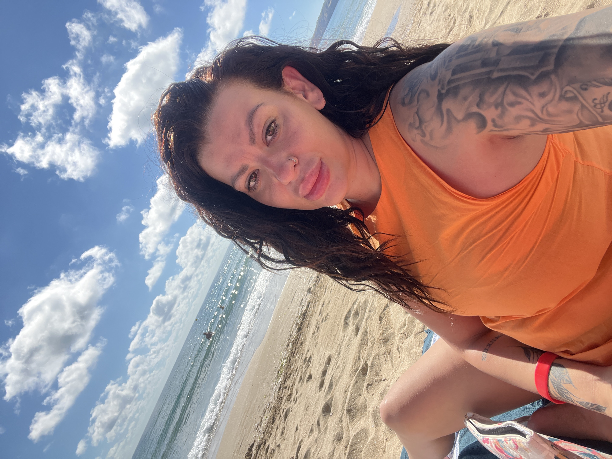 A woman with long, dark, wavy hair is sitting on a beach in Sozopol, Bulgaria, facing the camera. She is wearing an orange sleeveless top and has visible tattoos on her arm. Her legs are crossed, and a small patterned bag is visible next to her. The beach is sandy, with gentle waves lapping at the shore. The sky is blue with scattered white clouds, and the sea sparkles in the sunlight. Several people can be seen in the water in the background.