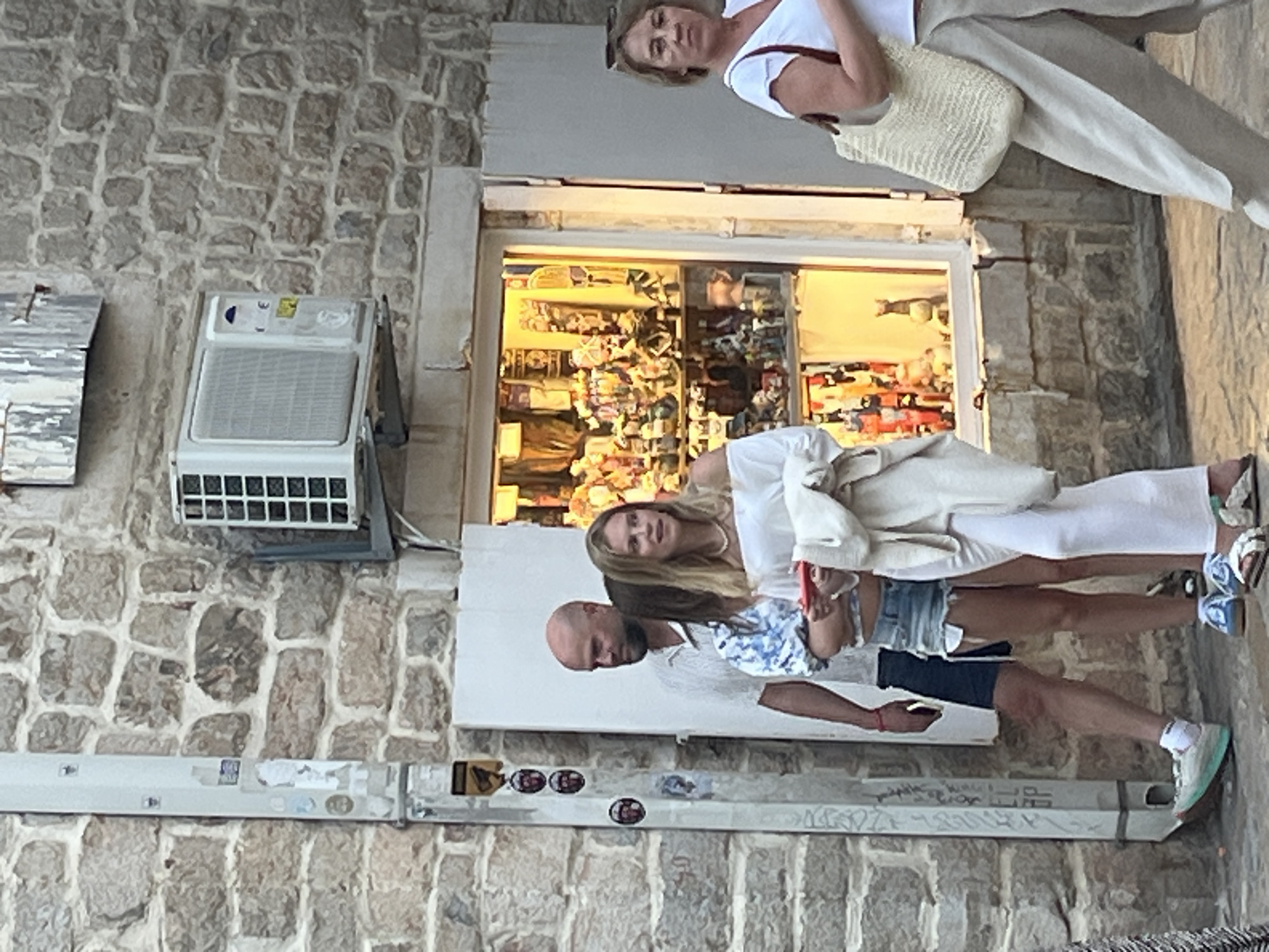 The image depicts a street scene in Budva, Montenegro. A man with a shaved head stands to the left, wearing a light-colored short-sleeved shirt and dark shorts. Beside him are two women. The woman in the middle wears a blue and white patterned top and denim shorts. The woman to her right is dressed in an off-the-shoulder white top and white pants, holding a red object. Further to the right, another woman, wearing a white top and light-colored pants, carries a straw bag.
In the background, a stone building features an open shop window displaying various items, and an air conditioning unit is mounted on the wall above. The street appears to be paved with cobblestones. The scene captures a moment of everyday life in the city.