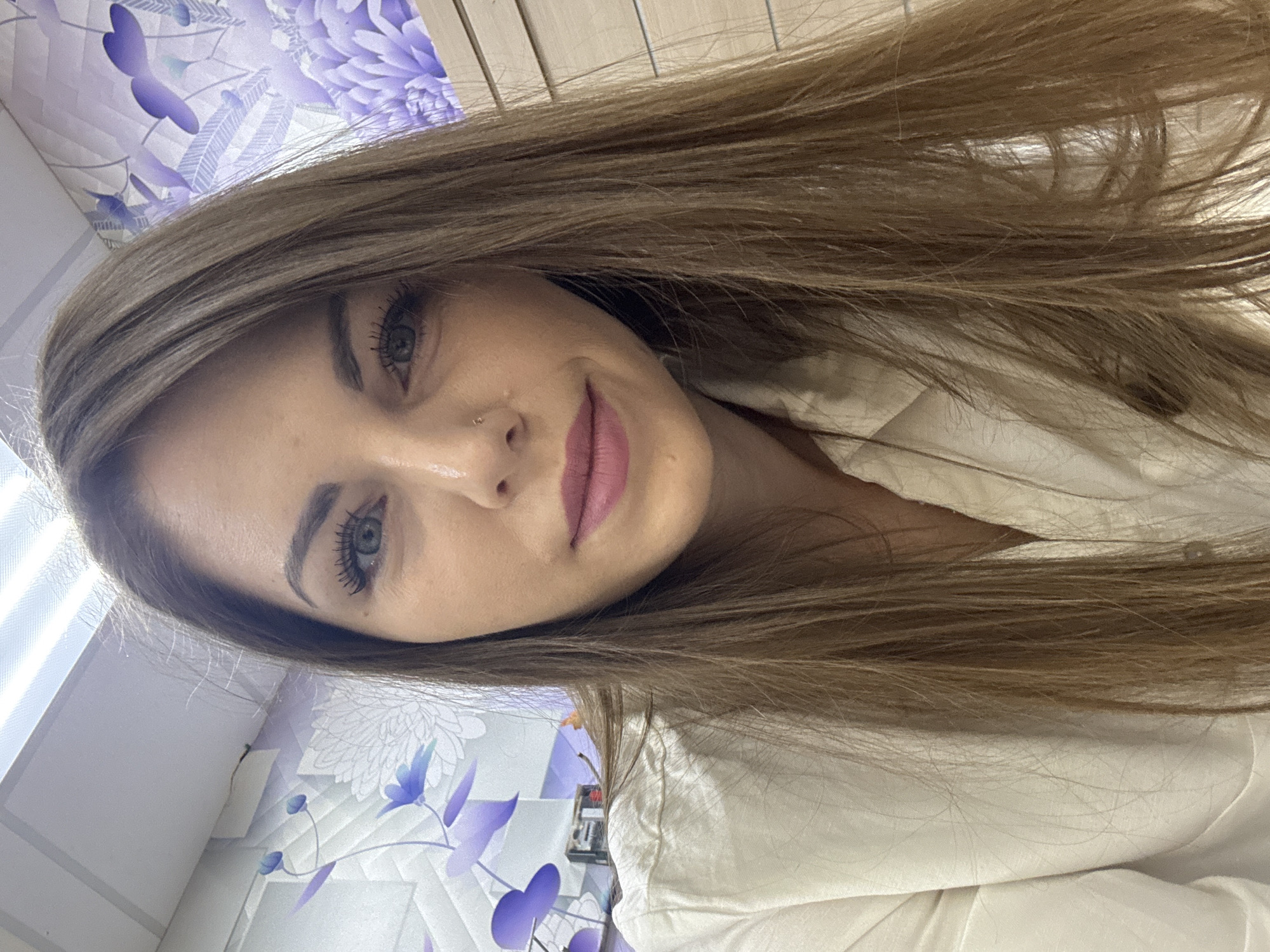 A close-up, selfie-style image shows a young woman with light brown hair. Her eyes are blue-grey, and she is wearing lipstick. She has a piercing in her nostril. She is wearing a white collared shirt. The background includes a wall with a light purple floral design and what appears to be a wooden venetian blind or a similar window covering. The location is potentially in Klin, Russia based on the provided context.