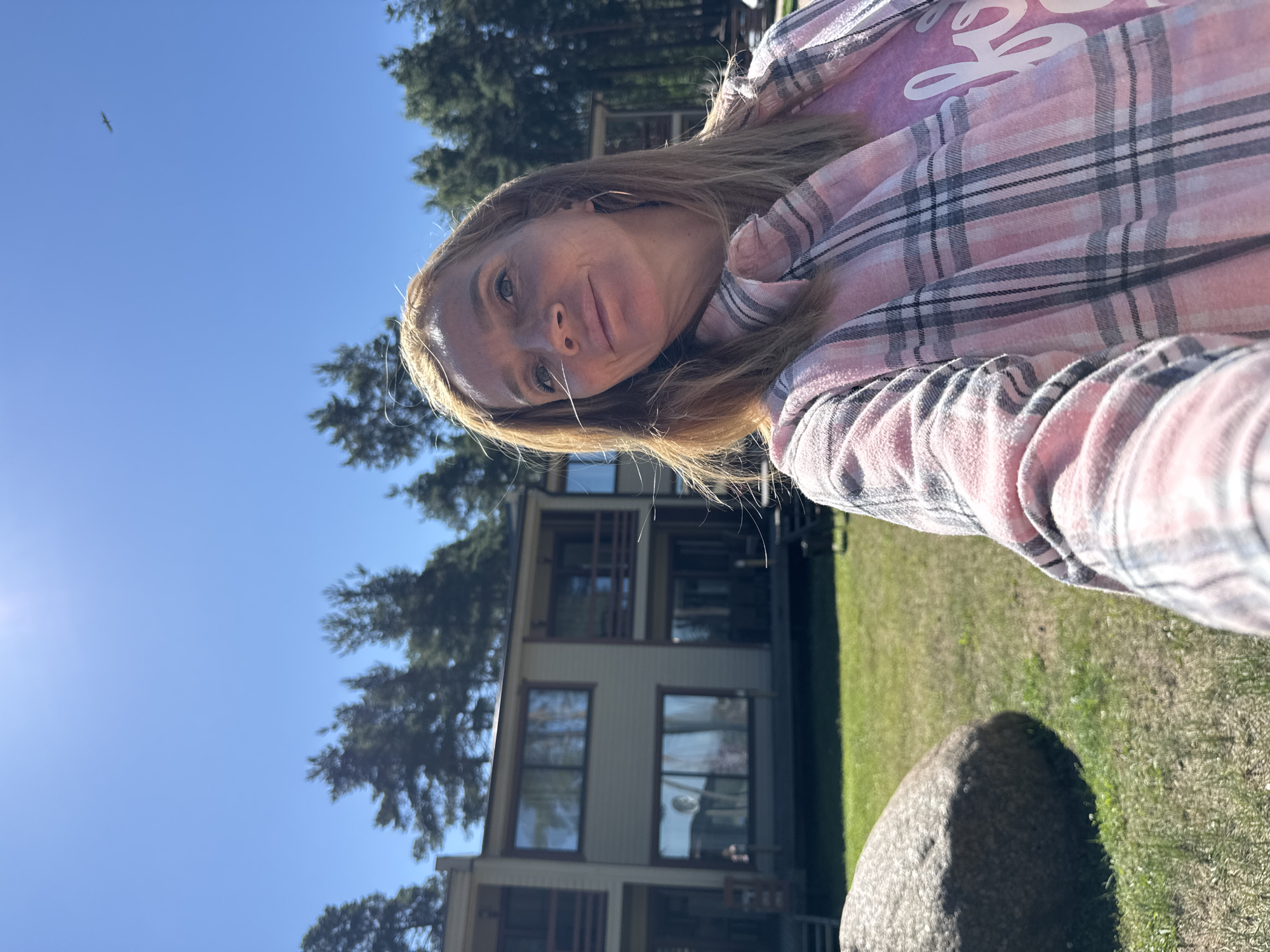 A woman with blonde hair is taking a selfie outdoors in a sunny environment. She is wearing a pink and gray plaid shirt over a pink t-shirt with white lettering. In the background, there are trees, a large rock, and a building that appears to be a multi-story residential structure with balconies and large windows. The sky is clear and blue, with a distant object that may be an airplane visible in the upper right portion of the frame. The setting suggests a location possibly within a suburban or resort area, given the presence of housing, natural elements, and an open outdoor space. The overall impression is of a casual outdoor moment on a clear day.