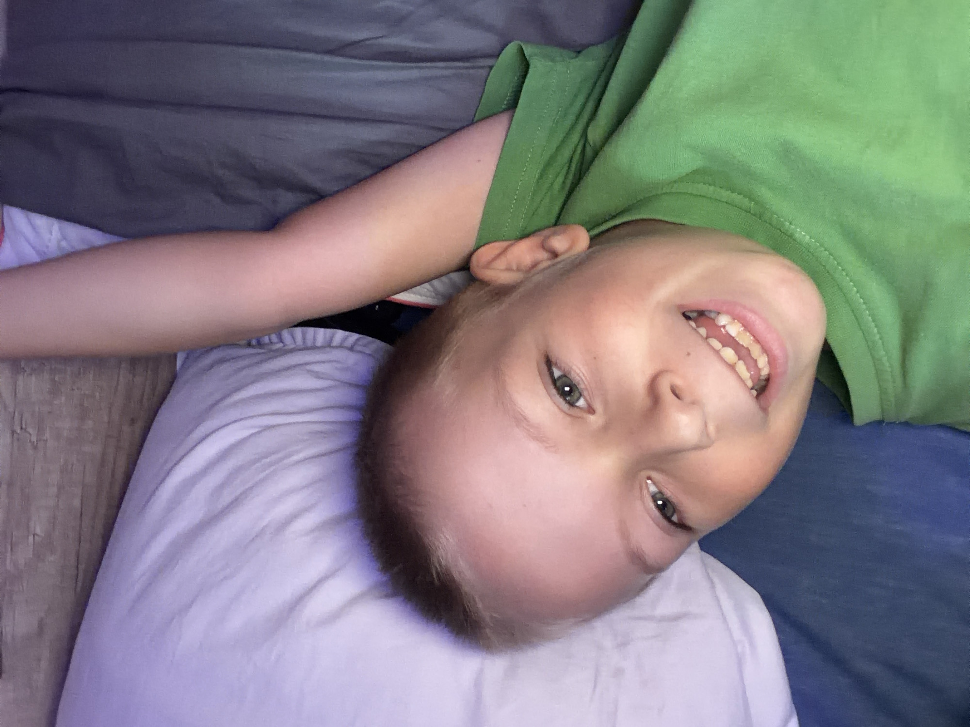 A young Caucasian boy with blonde hair and green eyes is lying on a bed. He is wearing a green t-shirt and his left arm is raised above his head. The boy is smiling and looking directly at the camera. The background consists of a white pillow, a grey duvet, and a wooden headboard. The image appears to have been taken indoors, likely in a bedroom. There is no visible indication of the specific location within Dolni Dabnik, Bulgaria.