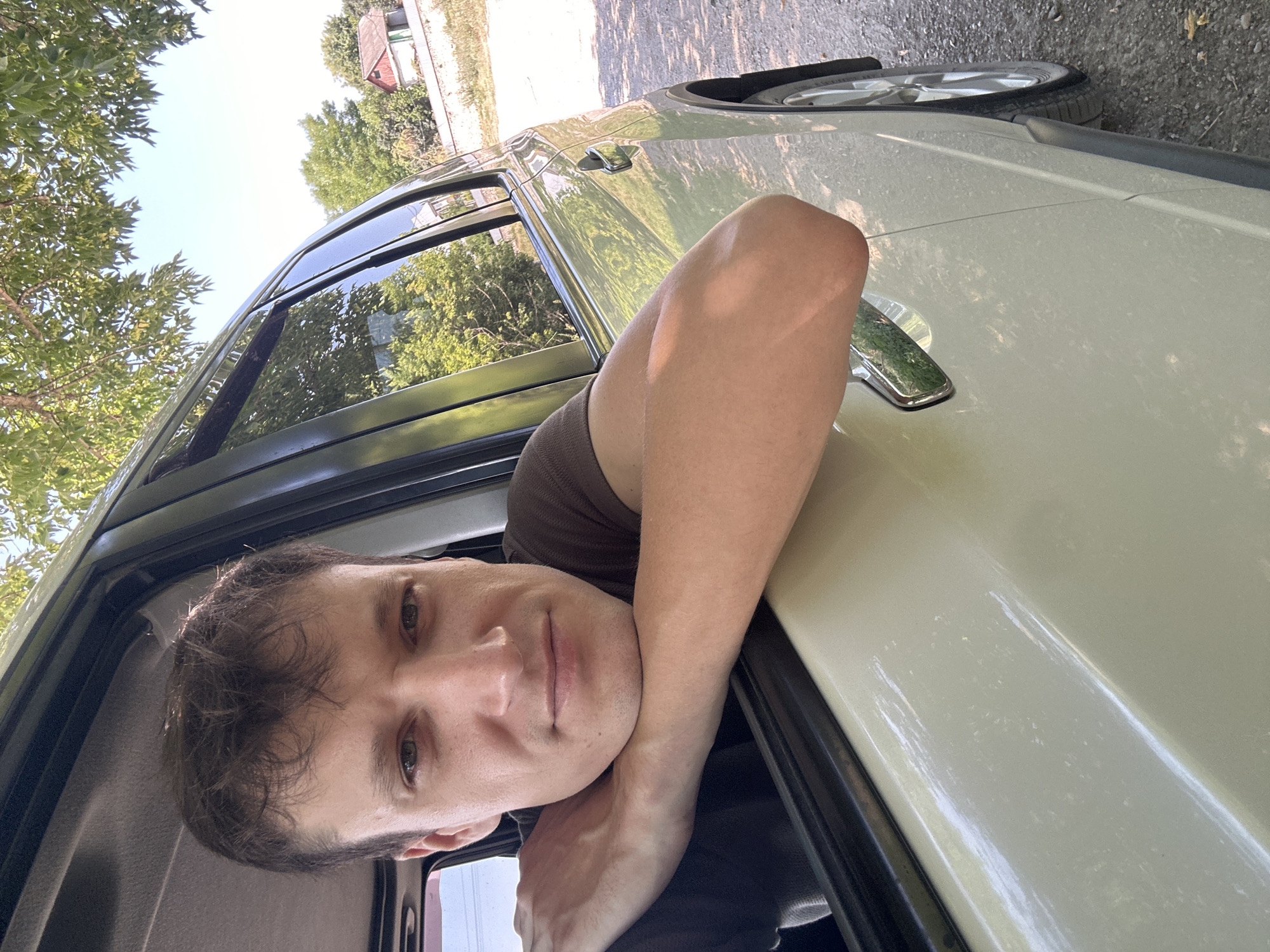In Makiv, Ukraine, a young man leans his arms out of the open window of a light-colored vehicle. He is wearing a brown t-shirt and his face is turned towards the camera, with his head resting on his crossed forearms. The car is parked on a gravel surface, with trees and vegetation visible in the background, including a house with a red roof. The sky is clear and blue, indicating a sunny day.
