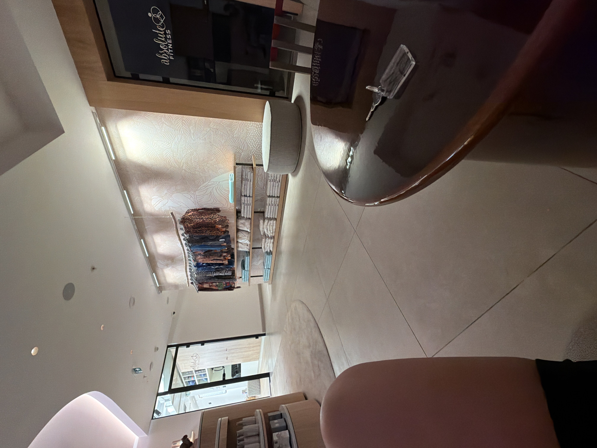 The image shows the interior of a retail space, likely a boutique or studio, with a focus on fitness apparel and accessories. The store features a modern design with clean lines and minimalist decor. In the foreground, a dark, polished round table is visible, with a clear plastic keychain and a key lying on its surface. To the left, a curved shelf unit displays rows of bottles and other products. Beyond this, a doorway leads to another area of the store, with shelving units visible inside. The main area of the store features a clothing rack displaying various patterned garments, hung neatly on hangers. Below the clothing rack, shelves are stacked with folded towels or linens. The background wall is decorated with a textured wallpaper featuring an organic, wavy pattern. A circular grey ottoman sits in the middle of the room. The lighting appears to be a mix of ambient and accent lighting, with LED strips illuminating the shelves and the textured wall. The flooring is tiled with large, light-colored tiles. The overall impression is one of a clean, organized, and stylish retail environment. This scene is located in Boreti, Montenegro.