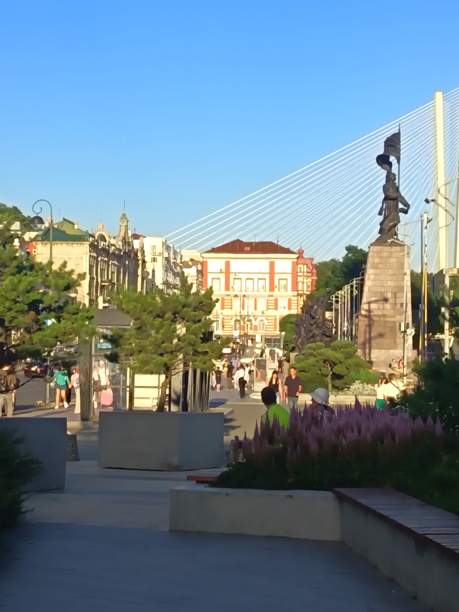 In an urban outdoor setting, under a clear blue sky, a broad, paved walkway is visible in the foreground. Several large, rectangular concrete planters are placed along the walkway. Beyond the planters, a bus stop shelter with glass panels stands on the left. To the right of the bus stop, a dense arrangement of purple-flowered plants creates a vibrant border. A wooden bench is situated in the lower right corner, extending along a planter.
In the midground, people are walking along the walkway. A statue of a figure holding a flag stands on a stone pedestal to the right, positioned in front of a suspension bridge with numerous white cables stretching across the sky. Behind the statue, several buildings with red and white facades are visible. A street with cars can be seen to the left of the bus stop.
In the background, trees and more urban architecture line the street. The scene appears to be bathed in the warm light of late afternoon or early evening.