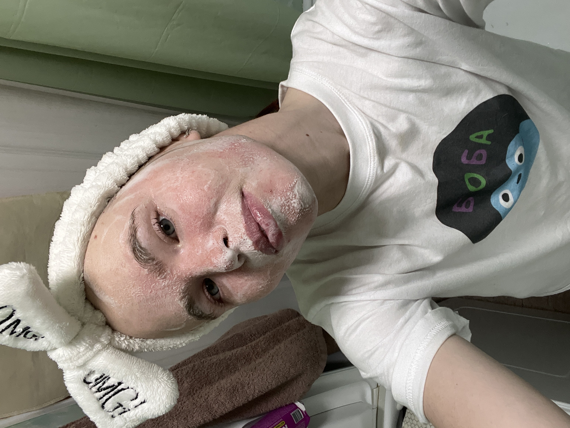 A person is pictured indoors, possibly a bathroom, in a selfie shot. The person has a white headband with bunny ears on their head. The headband has "OMG" embroidered on the ear. The person has a white foamy substance on their face, possibly a face mask or cleanser. They are wearing a white t-shirt with a stylized blob-shaped graphic on the front with the text "BOBA" in multiple colors and an image of a blue character with white eyes. A brown towel is draped over the edge of a surface and a purple container is visible behind it. The background is partially obscured but appears to be a light green and white painted wall. The location is Chaikovsky, Russia.