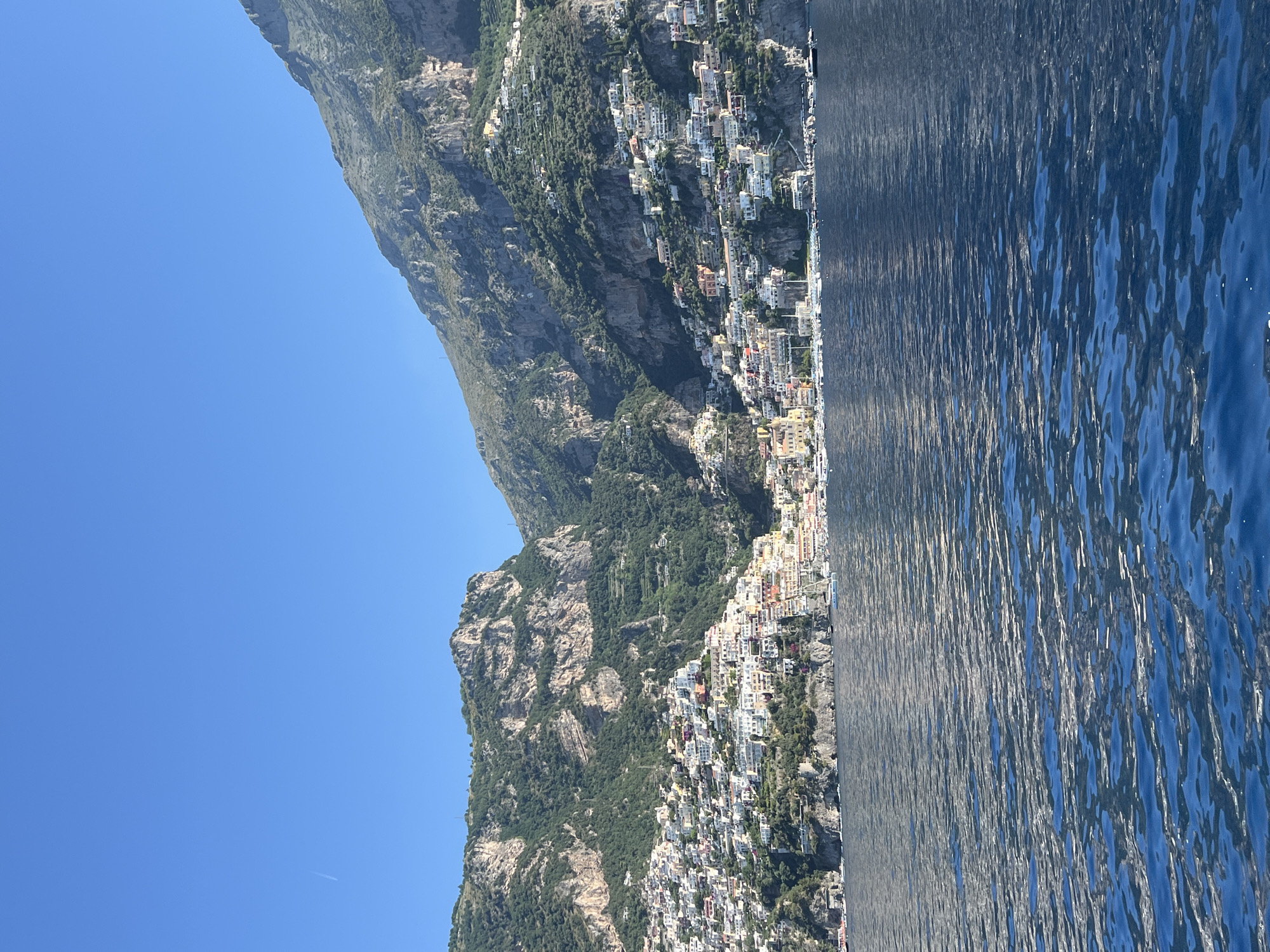 The image shows a coastal view of the town of Arienzo, Italy, from the water. The town is built on steep hillsides overlooking the sea, with colorful buildings clustered together. The mountains behind the town are covered in lush green vegetation and rocky outcrops. The sea in the foreground is a deep blue, with small waves reflecting the sunlight. A few boats are visible in the harbor area, and the sky is a clear, bright blue with a faint contrail from an airplane. The architecture is characteristic of coastal Italian towns, with pastel-colored houses, terracotta roofs, and narrow streets winding up the hillside.