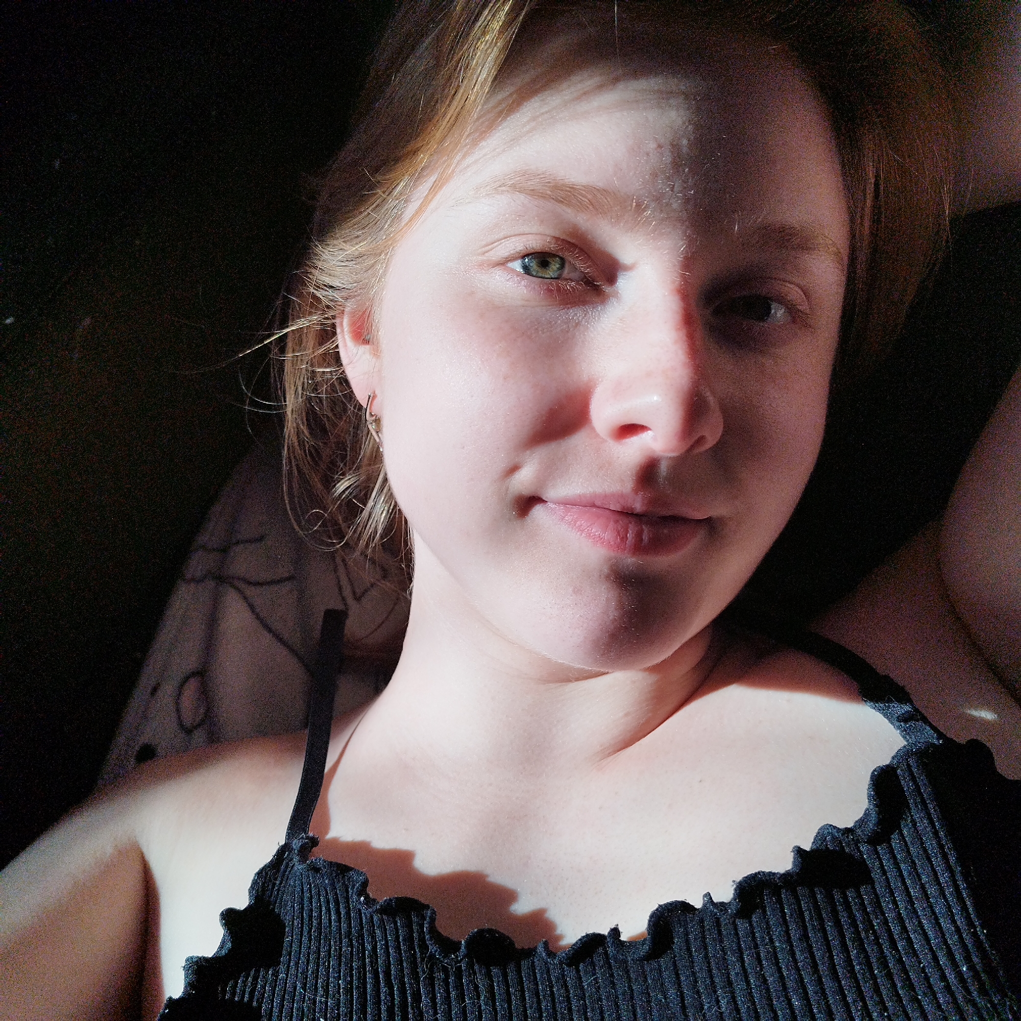 A close-up portrait of a young woman with fair skin and light brown or reddish-blonde hair, wearing a black ribbed tank top with a ruffled neckline. The lighting is dramatic, with strong contrasts between light and shadow across her face and body. Her eyes appear to be a greenish-hazel color. One can observe a small hoop earring in her left ear. The background is predominantly dark and out of focus. No details are discernable in order to provide information on Yekaterinburg, Russia.