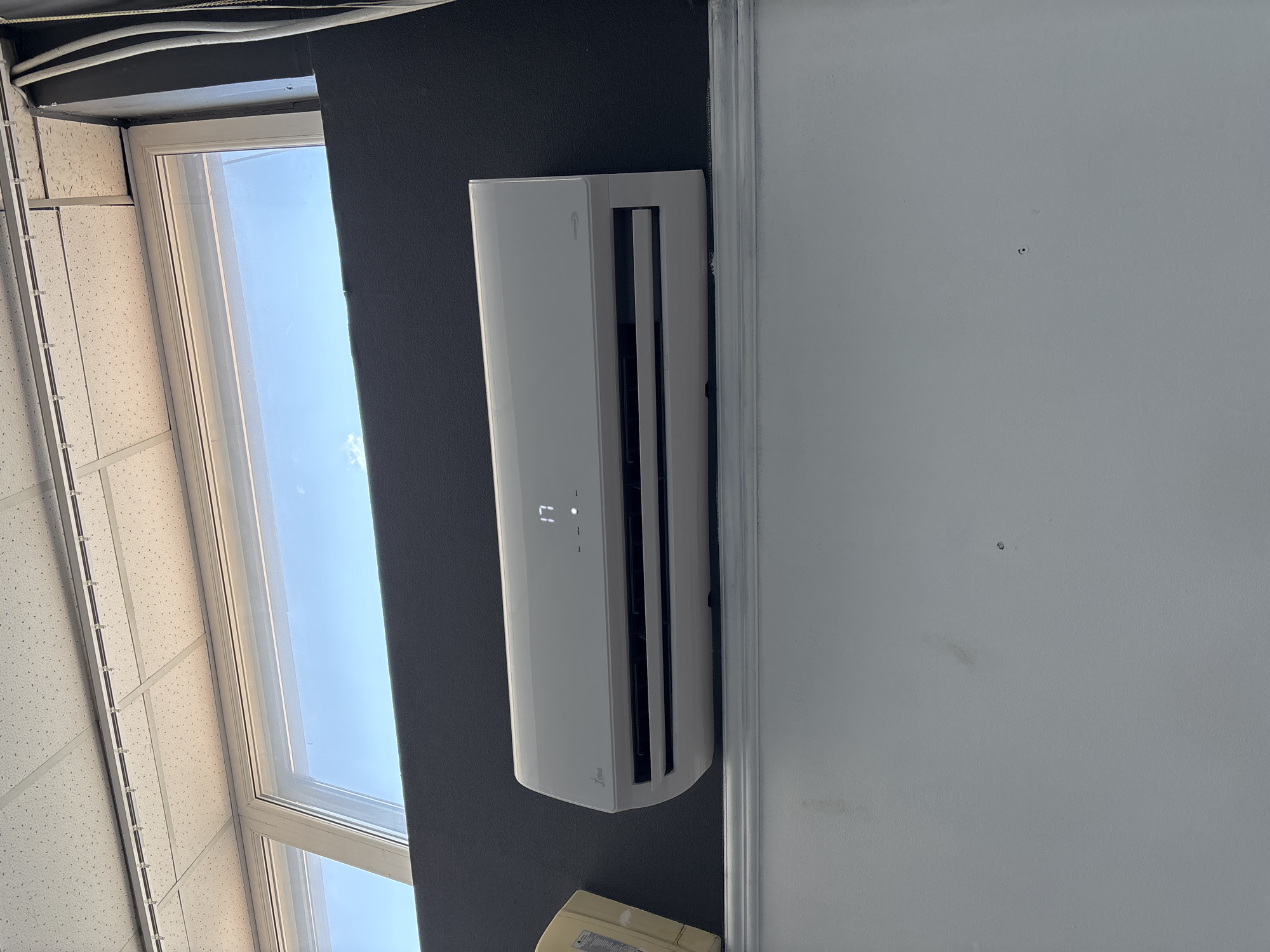 An indoor shot captures a white wall-mounted air conditioner unit with the temperature displayed as "17" on its LED screen. The unit is positioned centrally on the wall, below a large window that reveals a bright blue sky with a few small clouds. To the left of the air conditioner, a corner of a cardboard box is visible. The ceiling above features a grid of acoustic tiles with fluorescent light fixtures running along some of the grid lines. The lower portion of the image shows a white ledge or shelf beneath the air conditioner. The overall setting appears to be an indoor space with a view to the outdoors through the window, suggesting a room in a building.