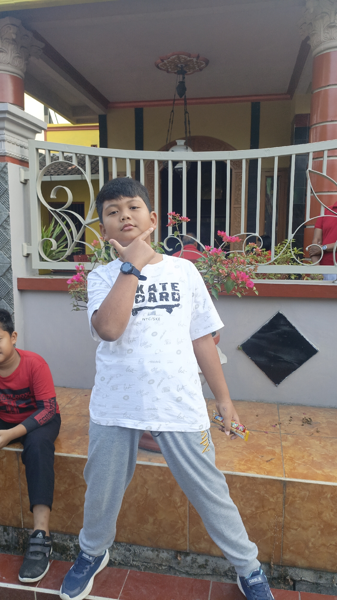 A young boy stands in front of a house with a white ornate railing and pink flowers. He is wearing a white t-shirt with black lettering, grey sweatpants, and blue sneakers. He has his right hand on his chin, and he is holding a candy in his left hand. The house is painted yellow, and there is a black diamond-shaped decoration on the wall. The boy is looking at the camera with a neutral expression. He is in the city of Salatiga, Indonesia.