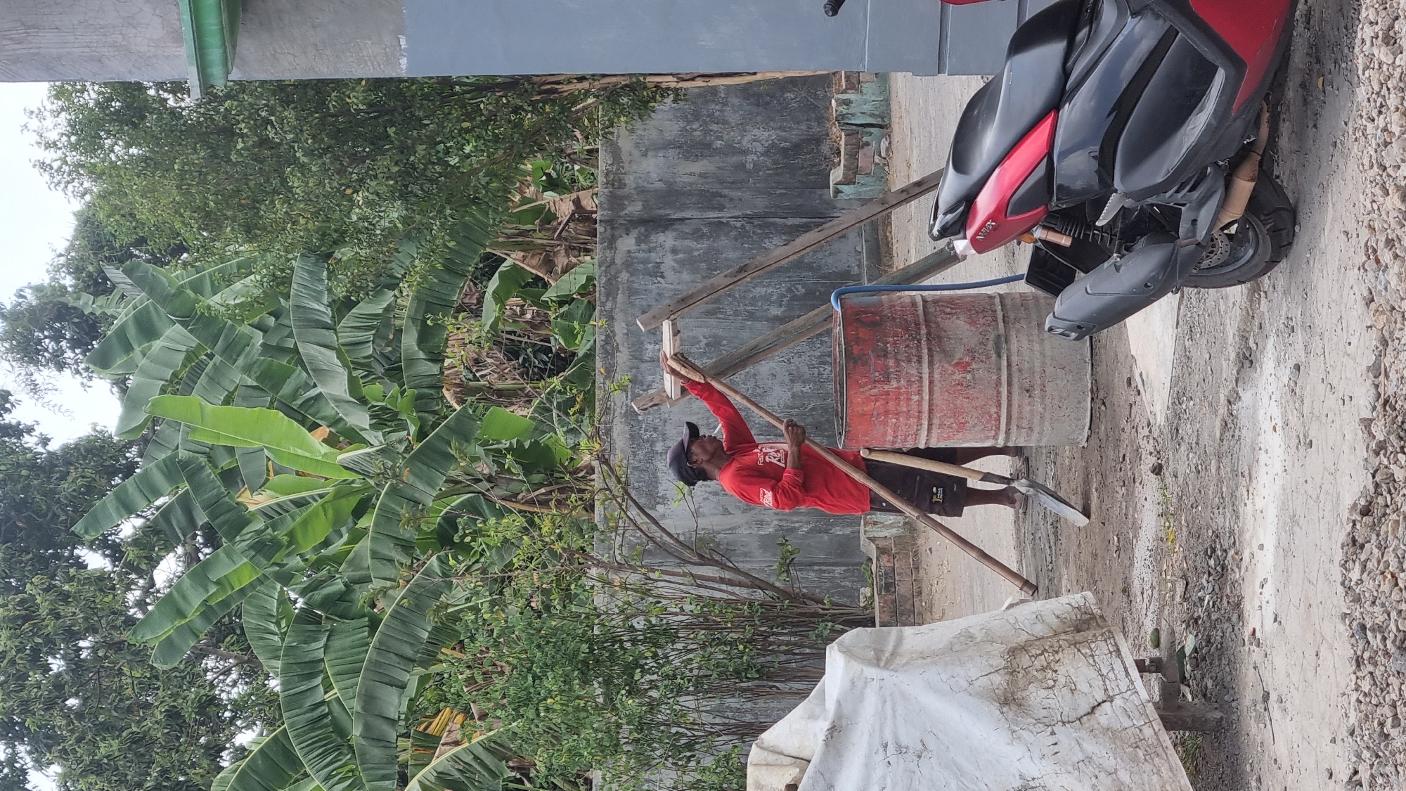 In Diwek, Indonesia, a person wearing a red long-sleeved shirt, dark shorts, and a black cap stands by a large red metal drum. The person is holding a long wooden pole and appears to be operating a manual mixer. A shovel is leaning against the drum. The scene is outdoors, with a concrete wall in the background and lush green banana trees and other vegetation. A white tarp covers an unseen object to the left, and a red and black motorcycle is parked to the right. The ground is a mixture of dirt, gravel, and some scattered white powdery residue.