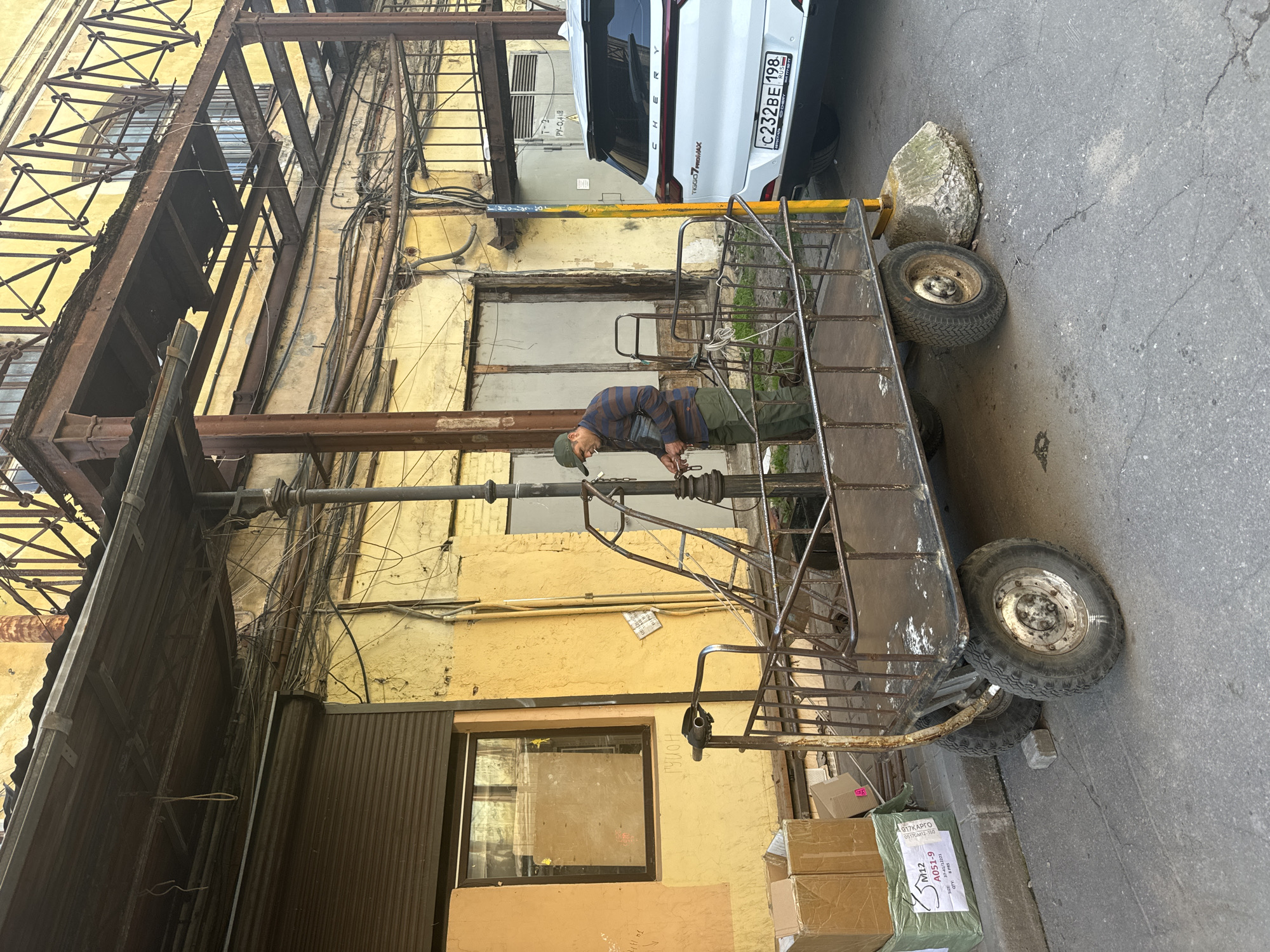 An outdoor scene in Saint Petersburg, Russia, shows a man standing next to a metal cart with two wheels and a wooden platform, appearing to tie a rope. The cart is positioned in an alleyway or service area next to a weathered yellow building with exposed metal beams and numerous wires overhead. A white Chery Tiggo 7 Pro Max SUV with a Russian license plate (C232BE 198) is parked to the right of the man and cart. Cardboard boxes are stacked on the left side of the frame. The ground is paved with asphalt, and a large, rough stone block is placed under one of the cart's wheels. The man is wearing a striped long-sleeved shirt, green pants, and a green baseball cap. He has a dark bag slung across his chest and appears to be smoking.