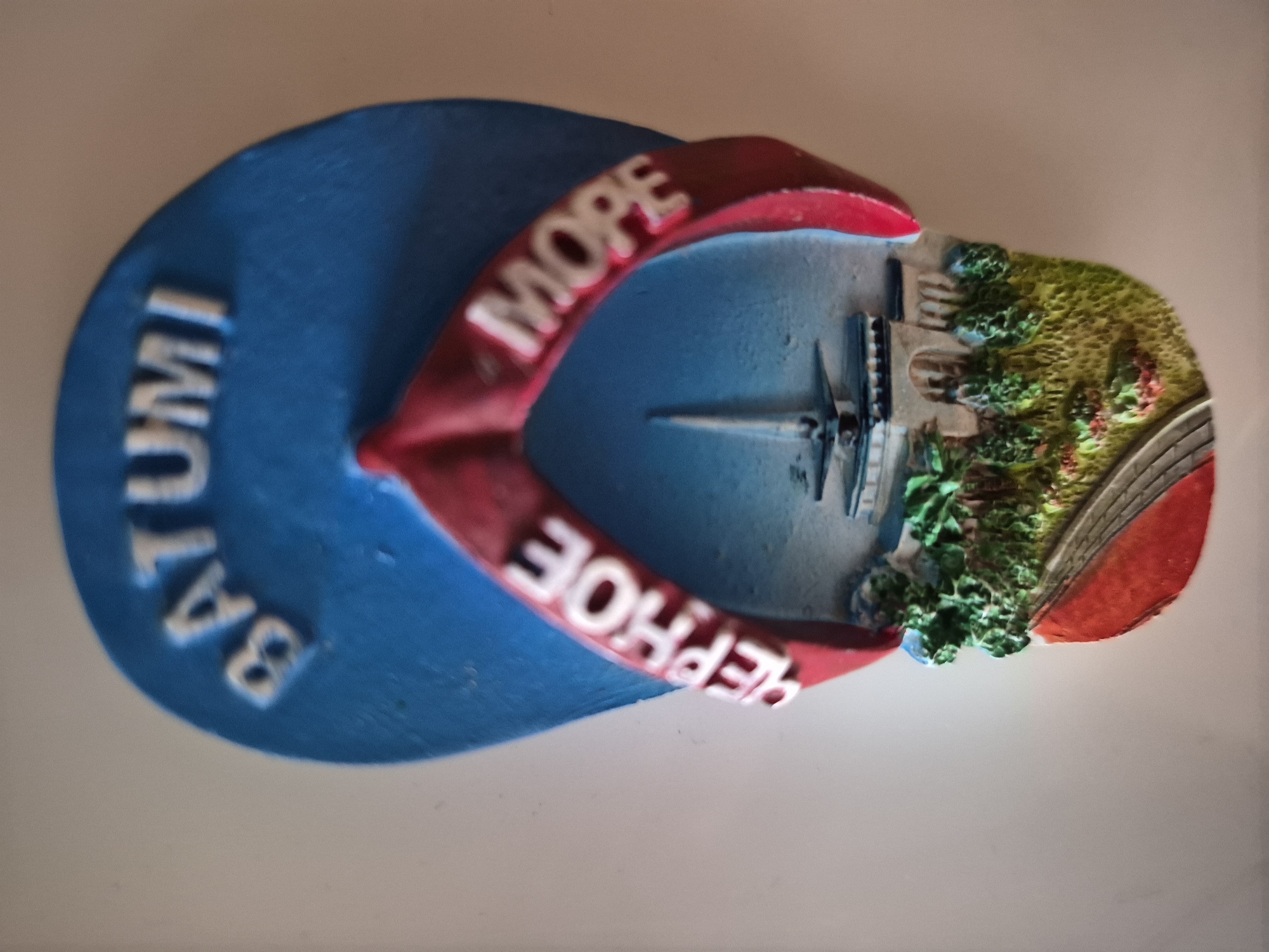 A souvenir magnet shaped like a flip-flop is displayed against a plain background. The top of the flip-flop is blue with raised white lettering that reads "BATUMI". The thong of the flip-flop is red and curves around a miniature scene. This scene depicts a building with a spire, surrounded by greenery and a red pathway. The words "ЧЕРНОЕ" and "МОPE" are also visible in raised white lettering on the red thong. The magnet is positioned at an angle, casting a shadow. There is no indication of the city of Vanadzor, Armenia, in the image.