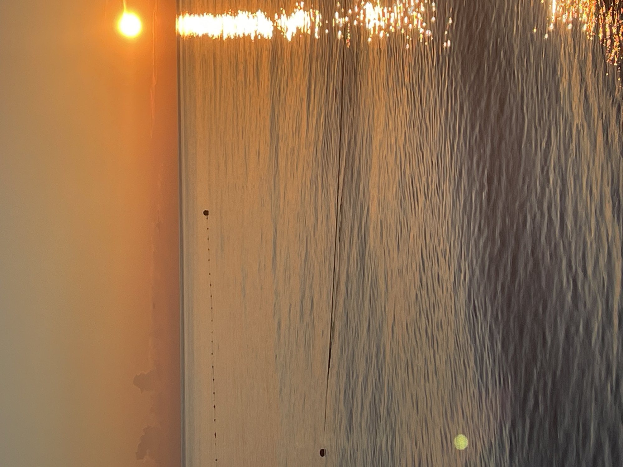 This is a vertical, medium shot image of a tranquil ocean at sunset. The sun is a bright orange orb setting on the right horizon, casting a shimmering reflection across the water. The sky is a gradient of warm oranges and yellows, with faint, wispy clouds near the horizon. In the foreground, the water is calm with gentle ripples. A line of buoys stretches across the middle ground, demarcating an area of the water. There is also a single, dark object, possibly another buoy, visible on the left side of the frame, closer to the viewer, and a faint green circular object in the lower left corner. The overall mood is peaceful and serene.