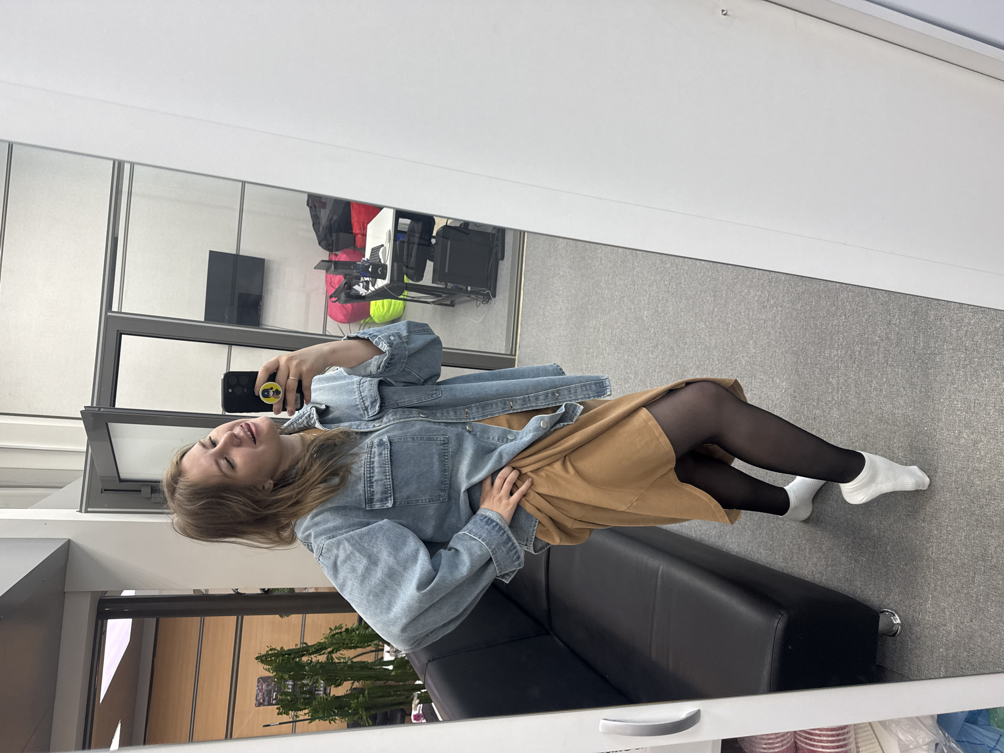 A young woman is taking a mirror selfie in what appears to be an office setting. She is wearing a light blue denim jacket, a tan skirt, black tights, and white socks. She is seated on a black leather-like sofa.
The reflection in the mirror shows a typical office interior, including a grey carpet, white walls and dividers, a TV or monitor mounted on a wall, and some miscellaneous office equipment. The presence of a plant near the window indicates a possible attempt to add greenery to the workspace.
Based on visual clues, the location may be Perm, Russia, but this cannot be confirmed.