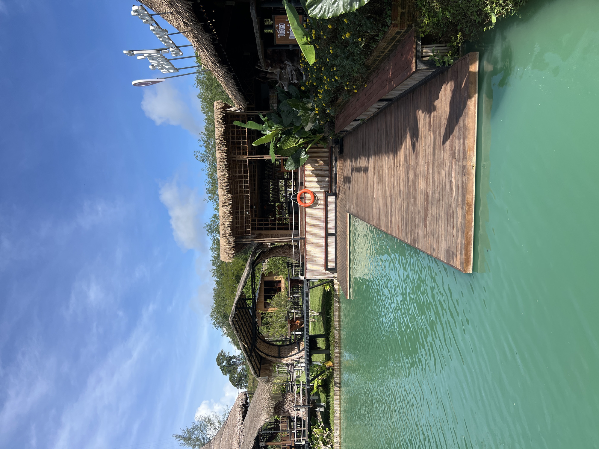 The image shows an outdoor scene with a body of turquoise water in the foreground, a wooden boardwalk extending into it, and several thatched-roof structures set back from the water. The structures appear to be part of a resort or restaurant, with some featuring open-air seating and bamboo detailing. Lush green vegetation, including large-leafed plants and yellow flowers, is visible around the buildings. A life preserver is hanging on one of the bamboo walls. The sky is blue with scattered white clouds, indicating fair weather. The location is identified as Tambon Pa Kae Bo Hin, Thailand. There are no people visible in the image.
