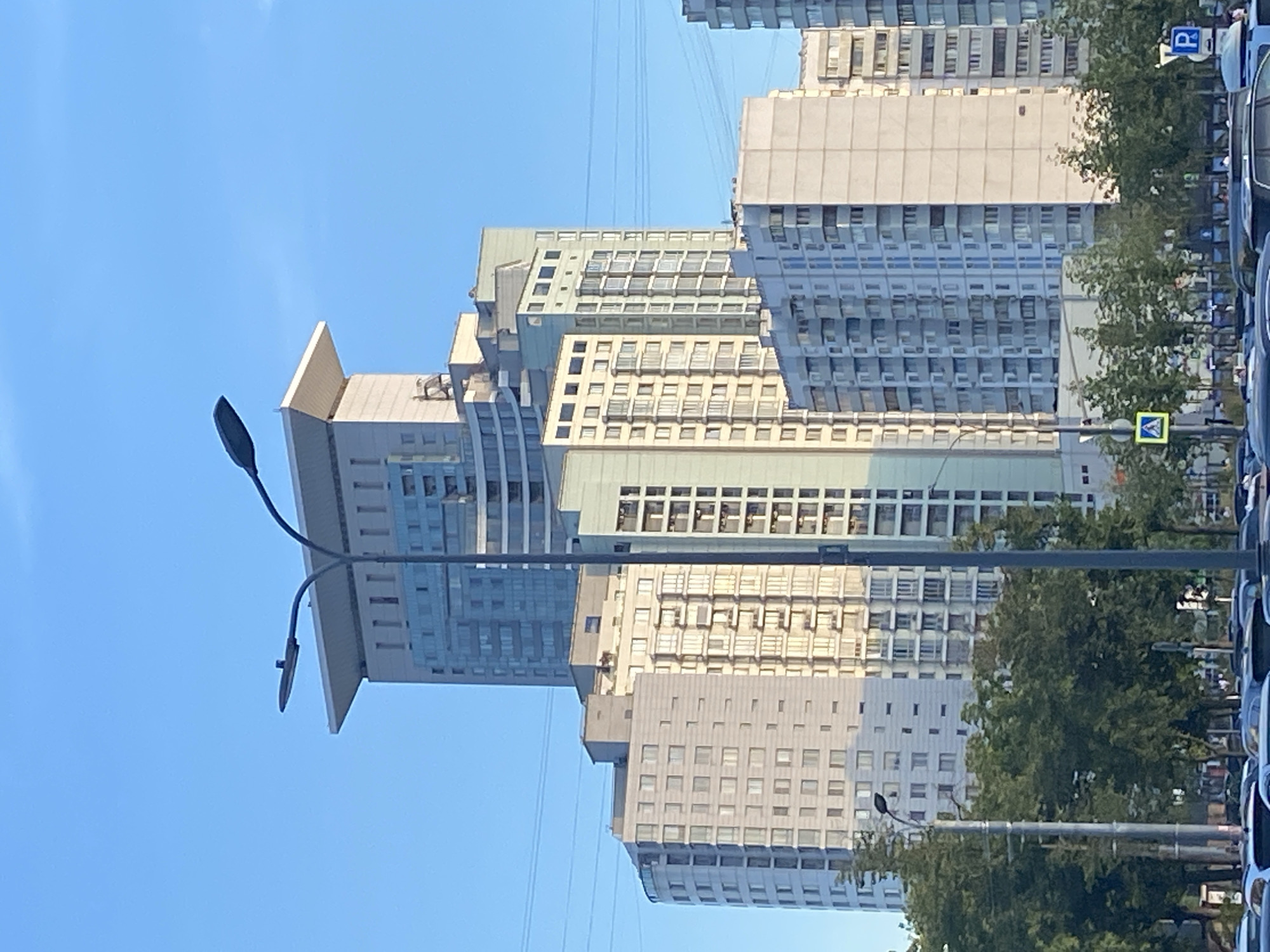 The image captures a wide-angle view of a modern urban street scene in Moscow, Russia, under a clear blue sky. Dominating the frame are several tall, multi-story residential buildings, characterized by their varied architectural styles and numerous windows. One prominent building towards the center of the image features a distinctive flat, cantilevered roof structure. In the foreground, a tall streetlamp with two fixtures stands centrally, and to its left, another streetlamp is partially visible. The street below is lined with trees, creating a green buffer between the buildings and the road. The road itself is occupied by numerous vehicles, suggesting active traffic, with a traffic light visible in the distance. Several road signs are also present, including a pedestrian crossing sign and a blue parking sign. The overall impression is one of a bustling residential area within a Russian metropolis, bathed in sunlight.