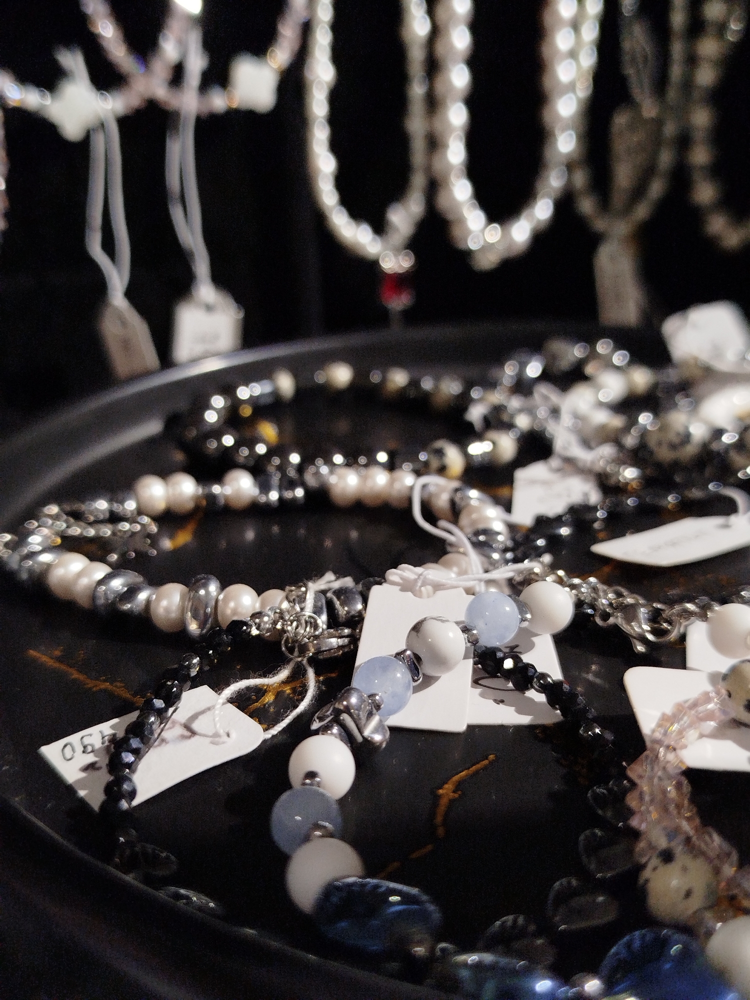 In an indoor setting, possibly a store or market stall, a variety of necklaces and bracelets are displayed on a dark, circular tray. The jewelry features a mix of beads, including white pearls, faceted black beads, blue beads, and metallic silver-toned beads. Some pieces are adorned with intricate silver chains. Several white tags are attached to the jewelry, some with partial price information visible, such as "06". The background is dark and out of focus, with blurred strings of beads hanging above. The overall lighting is dim, with highlights reflecting off the shiny surfaces of the beads and metal. There are no people visible in the image.