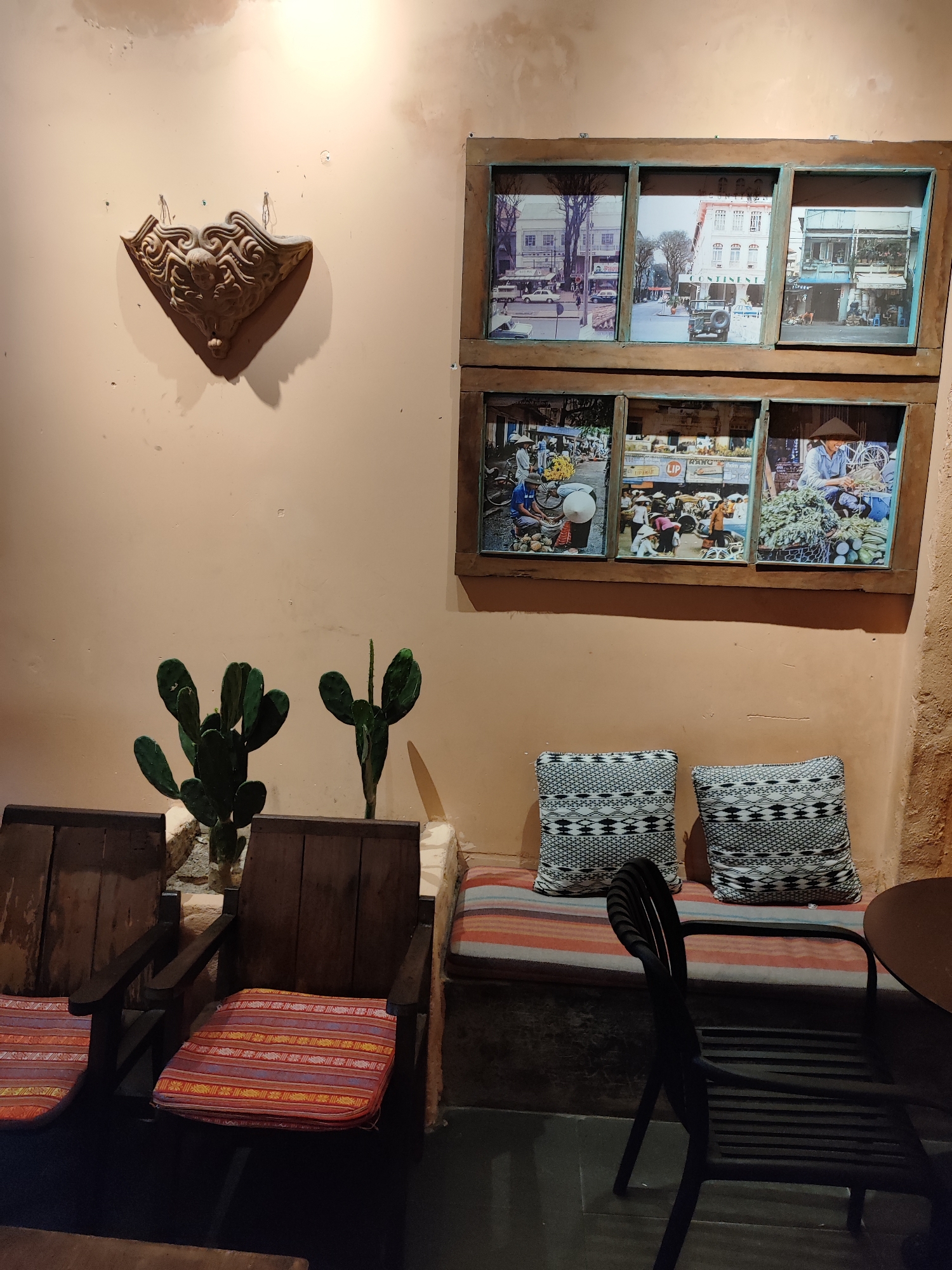 This image depicts a corner of a room, possibly a cafe or restaurant, in Da Nang, Vietnam. The walls are a muted peach color, with visible texture and some water stains near the ceiling. Mounted on the wall is an ornate, carved wooden decorative piece that resembles a shell or an elaborate crest. Below this, a large framed display contains six rectangular photographs arranged in a two-by-three grid. The top row of photographs shows street scenes with buildings and vehicles, suggesting an older era. The bottom row features images of market scenes with vendors and produce, indicative of local life.
In the foreground, two dark wooden armchairs with vibrantly striped cushions are positioned. Between the chairs and against the wall is a built-in bench with a striped cushion and two black and white patterned throw pillows. To the left of the chairs, two potted cactus plants are visible. To the right of the bench, a black metal chair is placed near a dark circular table. The flooring appears to be dark tile. The overall ambiance suggests a space that combines historical elements with comfortable seating, common in establishments aiming for a vintage or culturally inspired aesthetic.