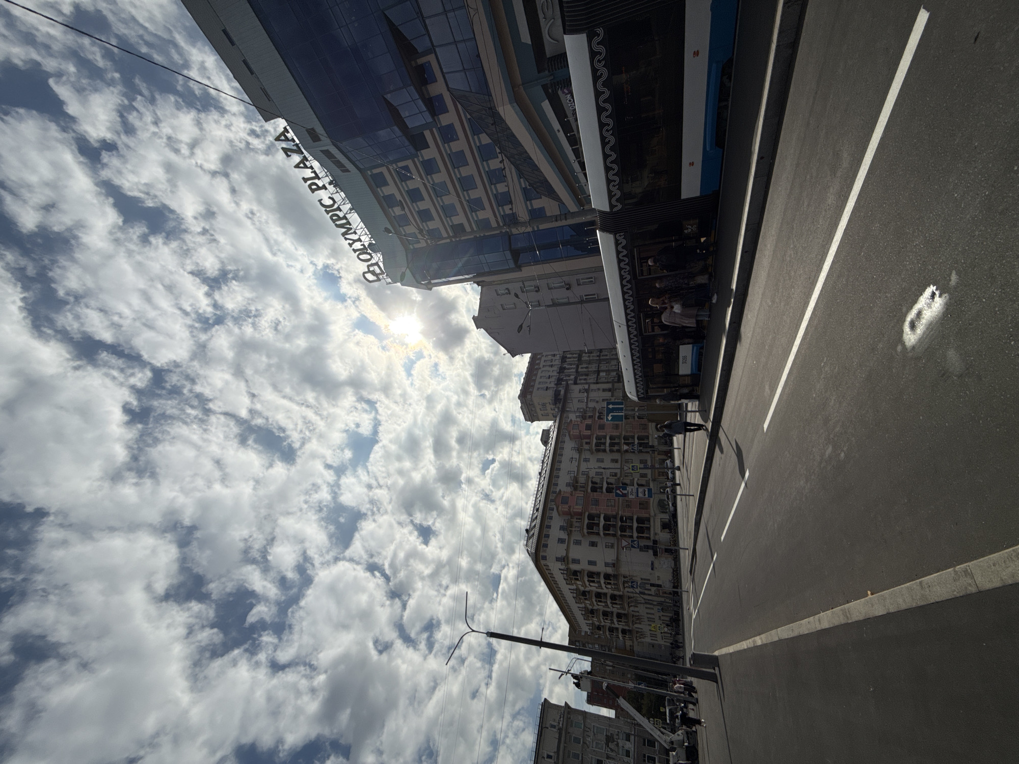 This is a street-level, wide-angle shot of a city street in Moscow, Russia, on a cloudy day. The sky is filled with dramatic, textured clouds with the sun partially visible through them, casting a bright light. The scene features several buildings, a street, a tram, and a few pedestrians.
On the right side of the image, a modern, multi-story building with a glass facade and the prominent signage "OLYMPIC PLAZA" stands tall. Below this building, a tram is stopped, with several people waiting to board or disembark. The tram is white and blue with decorative black wavy patterns along its upper edge. Some people are visible inside the tram.
To the left of the Olympic Plaza building, there are older, residential buildings with multiple balconies and architectural details in earthy tones, some accented with pink. A street lamp with an unusual shape stands in the middle ground, and a construction crane is visible in the distance on the far left.
The street itself is paved with asphalt and marked with white lines. A sidewalk runs along the left side of the street, and further to the left, more buildings are visible. Pedestrians are scattered across the scene, walking on sidewalks or waiting at what appears to be a tram stop. The overall impression is of an active urban environment under a dynamic sky.