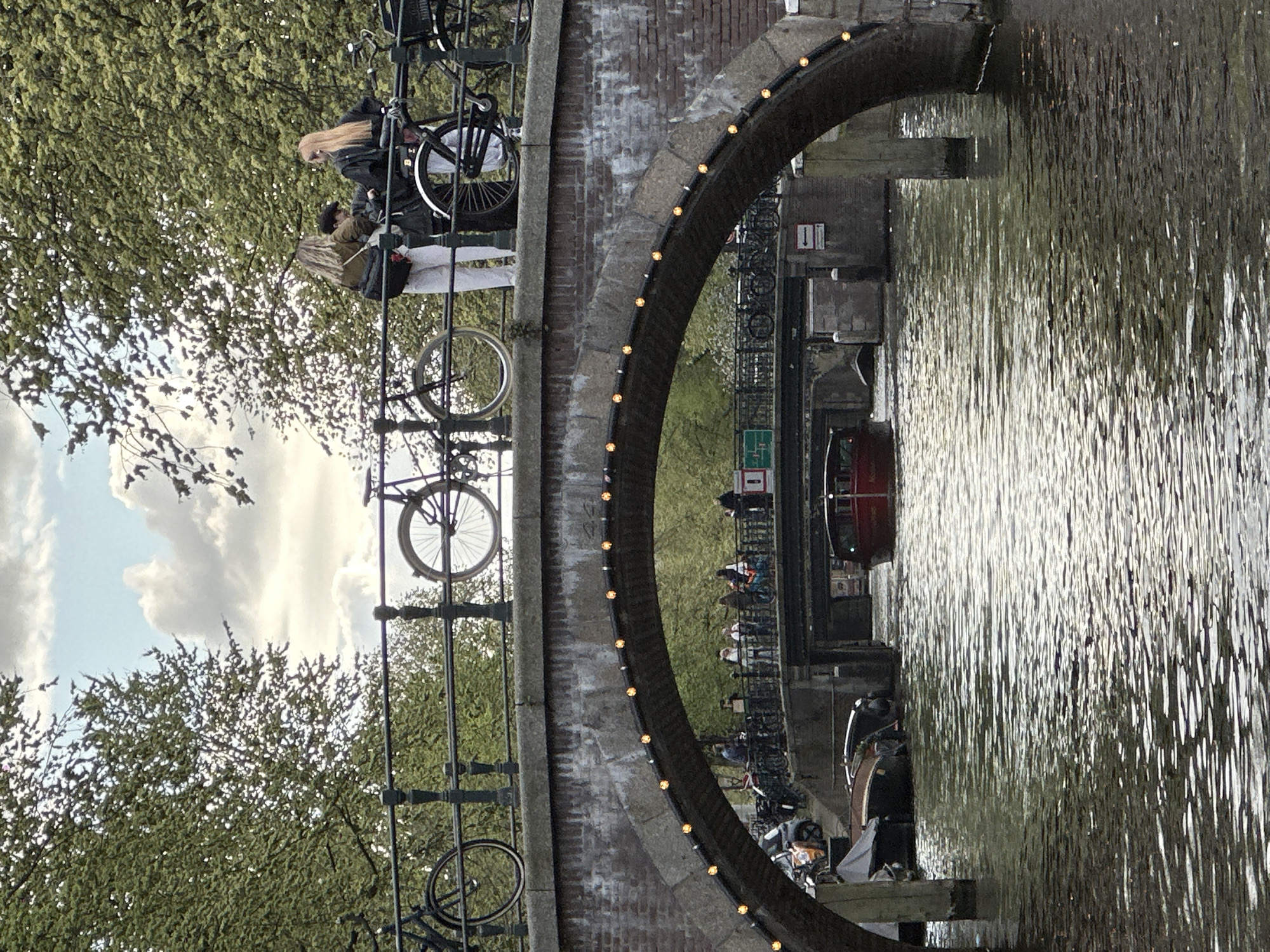 This media file captures a lively canal scene in Amsterdam, Netherlands, oriented incorrectly as if rotated 90 degrees clockwise. When viewed correctly, it depicts a daytime setting, possibly late afternoon, under a partly cloudy sky.
In the foreground, a sturdy stone or brick arch bridge gracefully spans a bustling canal. The bridge's arch is adorned with a decorative string of warm-toned lights. Numerous bicycles, characteristic of Amsterdam, are parked along the bridge's railing. On the bridge, several people are visible, including two individuals prominent on the upper left side: one with long blonde hair and another with darker hair, standing near parked bikes.
The canal water shimmers with bright reflections of the sky and surrounding foliage. A red and dark-hulled boat is seen navigating under the bridge, with other smaller boats and people visible further along the canal banks. Lush green trees and foliage dominate the scene, framing the canal and extending upwards towards a sky dotted with white clouds.
To the left of the bridge's arch, by the canal, a small sign featuring two red squares, each containing the white number "11," is visible. The image conveys a sense of peaceful urban activity, showcasing the distinctive elements of Amsterdam's iconic canals and bike culture.