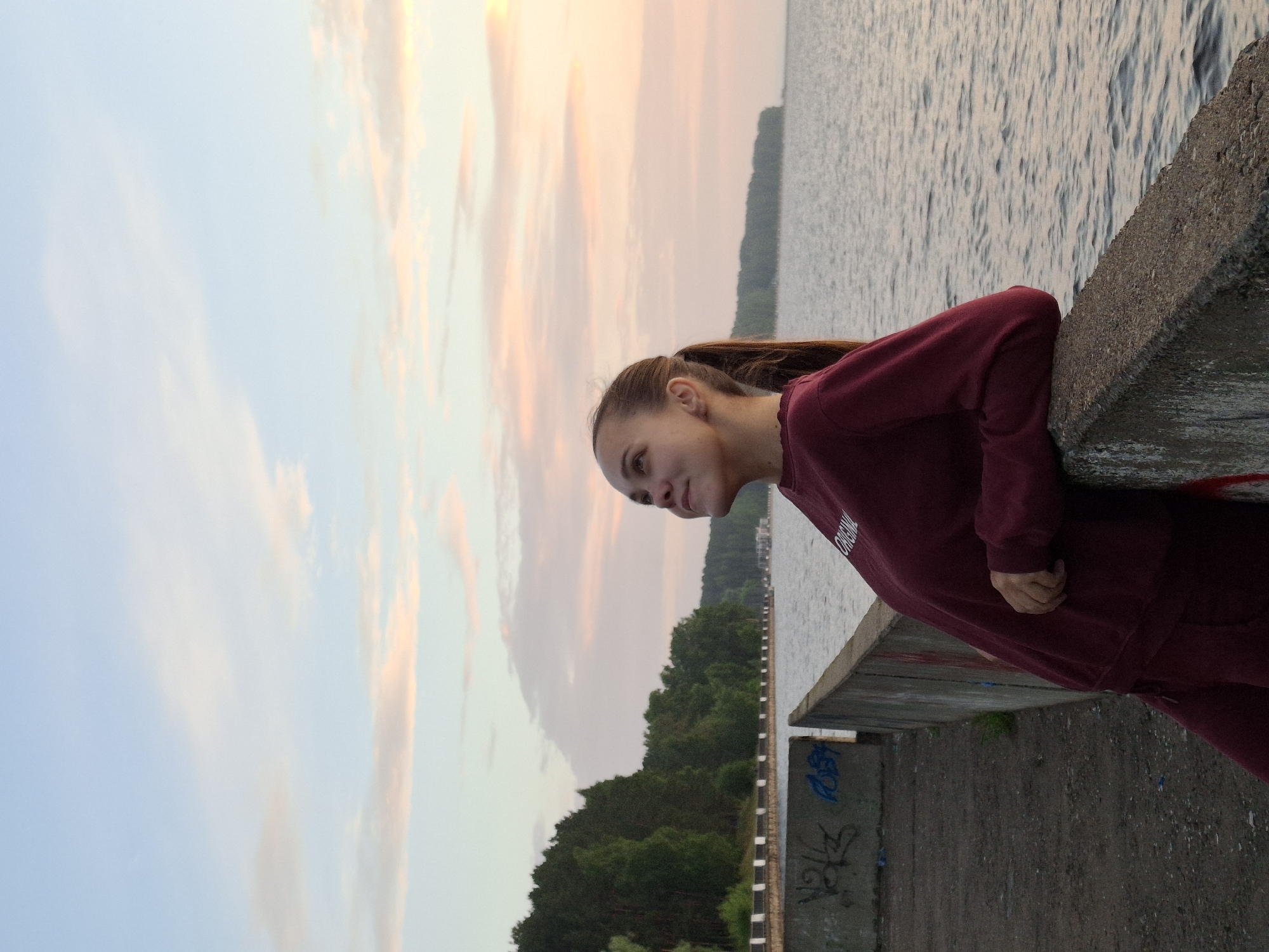 A young woman with her hair in a ponytail stands on a concrete embankment next to a body of water, looking up at a pastel-colored sky at sunset. She is wearing a burgundy sweatshirt and matching sweatpants. The embankment appears to be part of a promenade or waterfront area in Chaikovsky, Russia, evidenced by the trees and shoreline visible across the water. The sky is filled with soft clouds in shades of pink, orange, and blue. The water is calm with gentle ripples. Graffiti is visible on the concrete structures.