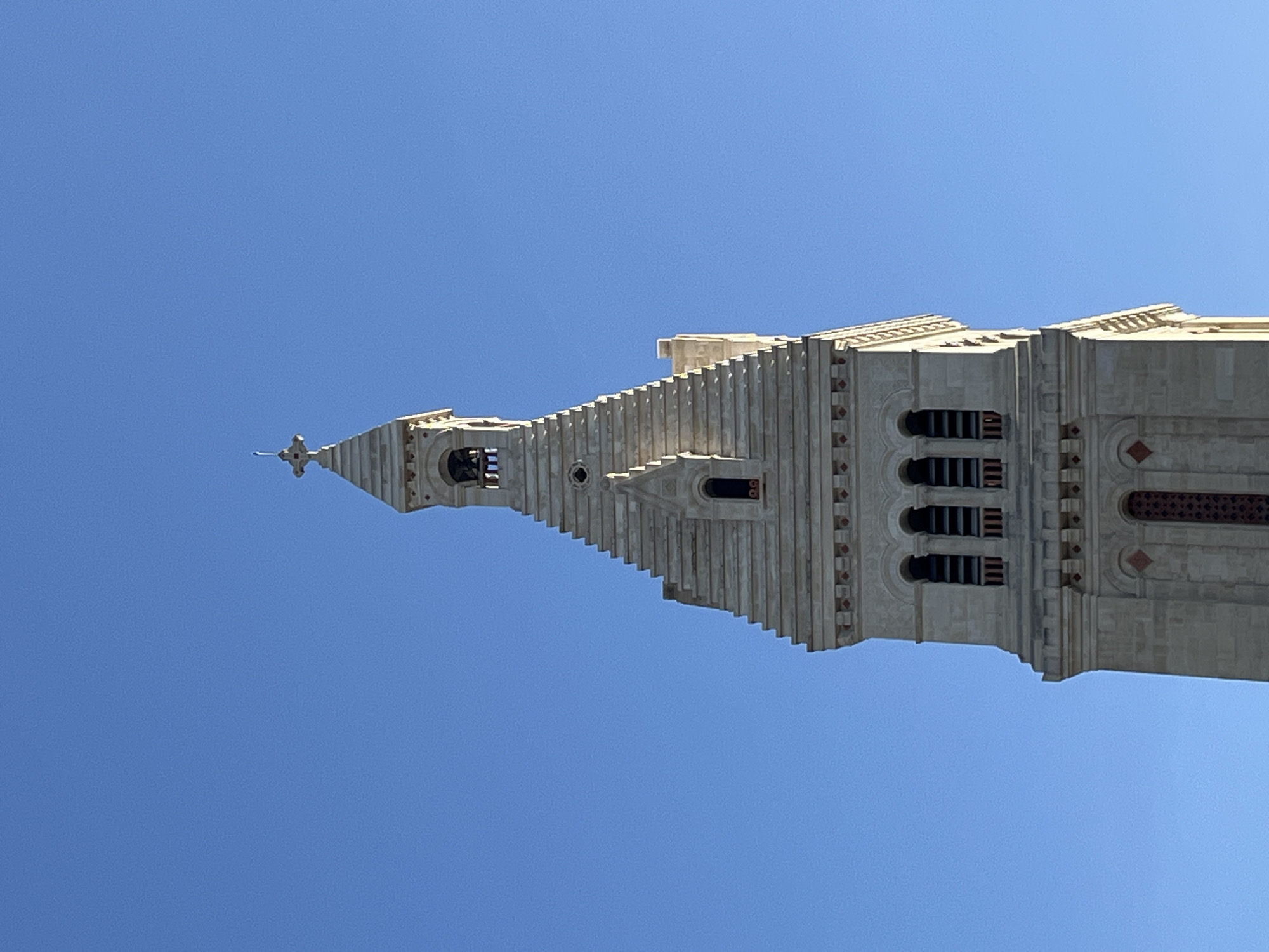 The image shows the bell tower of the Sacré-Cœur Basilica in Paris, France, against a clear blue sky. The tower is constructed of light-colored stone and features a stepped pyramidal roof, topped with a cross. Several arched windows are visible, with three larger arched openings on one level, likely housing bells. Below these, there are smaller, narrower windows and decorative stonework. The bell tower is a prominent religious architectural structure.